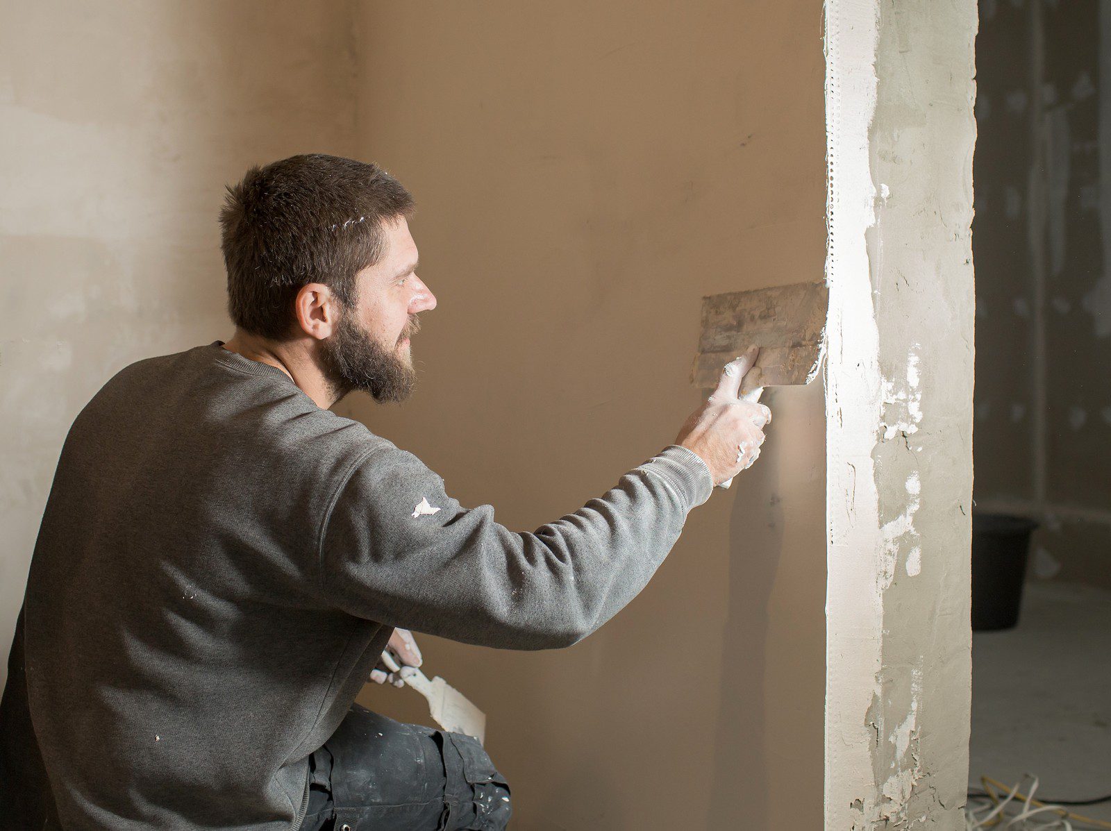 The image shows a man engaged in some type of construction or repair work, specifically applying plaster or joint compound to a wall. He is using a hawk, which is holding the plaster, and a trowel to smooth the material onto the wall. The man is wearing casual work clothes which show signs of wear from his labour, and he appears to be focused on his task. The room appears to be unfinished, with bare walls and some construction materials or debris on the floor.