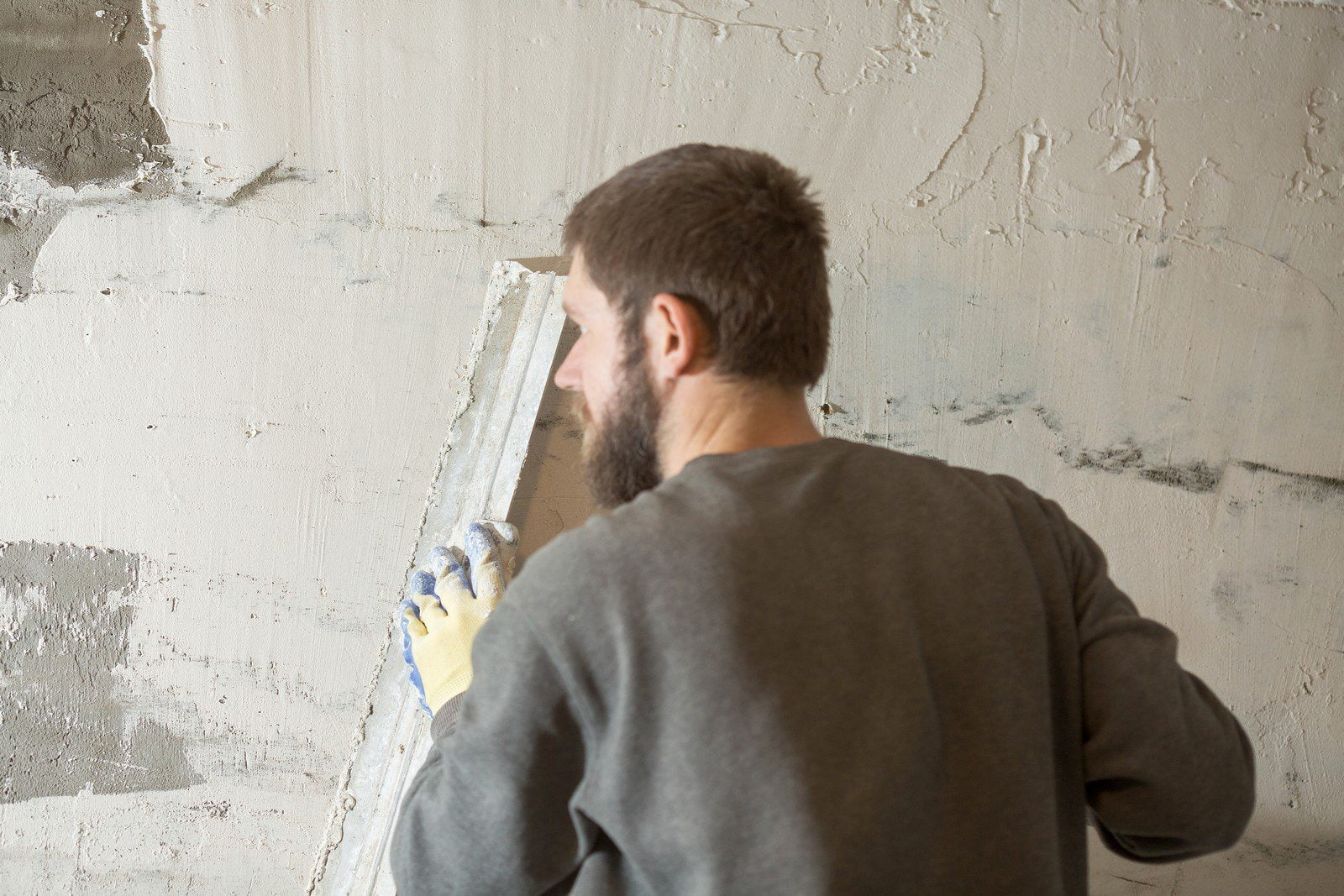 The image shows a person from the back and side profile, applying a substance to a wall with a trowel. The person is likely a construction worker or a DIY enthusiast engaged in plastering or mudding a wall, which is part of the process of finishing walls in construction. The wall appears to be partially covered with the plaster or compound, and there are areas of uneven application suggesting that the work is in progress. The worker is wearing a long-sleeve shirt and protective gloves, which is common for this type of work to protect the skin and improve grip.