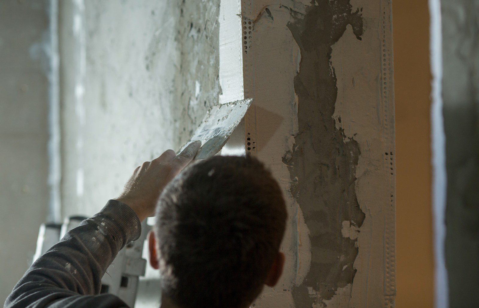 This image shows a person applying plaster or drywall compound to a wall using a trowel. The person is likely engaged in construction or renovation work, specifically in the process of wall finishing or repair. The wall has patches where the plaster has been applied over what appears to be strips of joint tape or mesh, used to strengthen and smooth the joins between pieces of drywall or to repair cracks. The lighting suggests an indoor setting and the presence of construction or renovation activity.