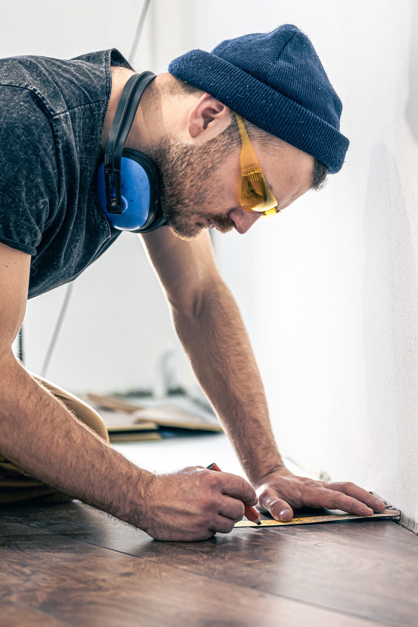 The image depicts a male worker in the process of installing laminate flooring. The man is positioned crouched down on a partially finished floor inside a room that appears to be under renovation. He is surrounded by flat, rectangular pieces of laminate flooring, some of which have already been laid down while others are yet to be installed. The worker is wearing a casual long-sleeve shirt, work pants, and sturdy shoes appropriate for construction activities. His attention is focused on an individual laminate plank he is handling, which he is carefully fitting into place with precision. The room's walls are plain and unadorned, suggesting ongoing construction or refurbishment. In the background, tools and materials are scattered, including a measuring tape and a toolbox, indicating an active work environment. Natural light filters into the room from an unseen window, illuminating the space with a soft, clean light.