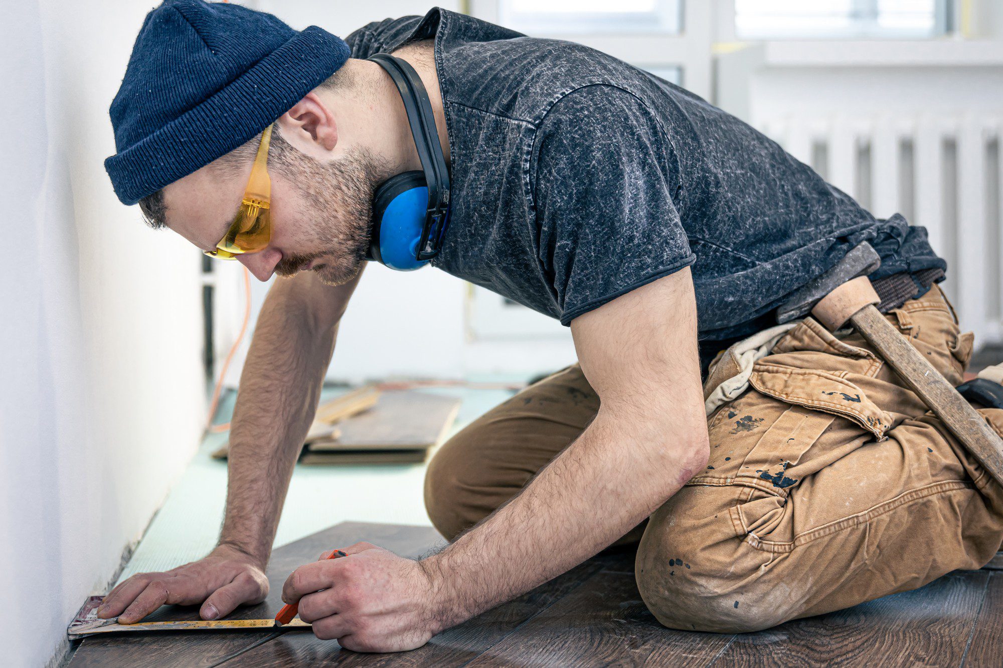 This image shows a person kneeling on a wooden floor while performing some kind of work. The person is wearing a beanie, safety glasses, a short-sleeve t-shirt, ear defenders or ear protection headphones, and heavy-duty work pants. It appears that the individual is marking or measuring something on a piece of material—possibly wood, as might be done during a flooring installation or carpentry work. We can see a ruler or tape measure and possibly a pencil or marking tool in hand. The environment suggests a work or construction site indoors as the background looks like a partially finished room with an unpainted wall and tools or materials strewn about.