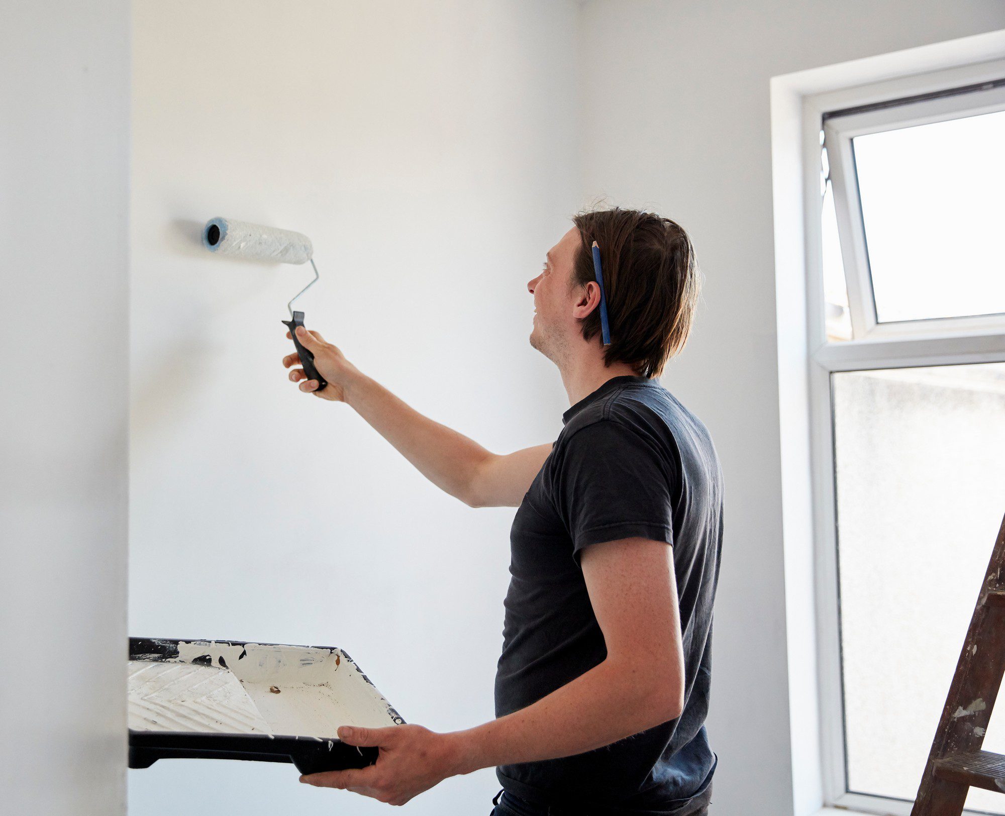 This image shows a person painting a wall with a roller. The individual is holding a paint roller in one hand and a paint tray in the other. The wall appears to be in the process of being painted, as evidenced by the patch of fresh paint where the roller is being applied. Next to the person is a small open window providing natural light to the room. The person is standing on a wooden ladder, indicating they are working at an elevated height to reach the upper portions of the wall. The environment suggests a home-improvement or renovation setting.