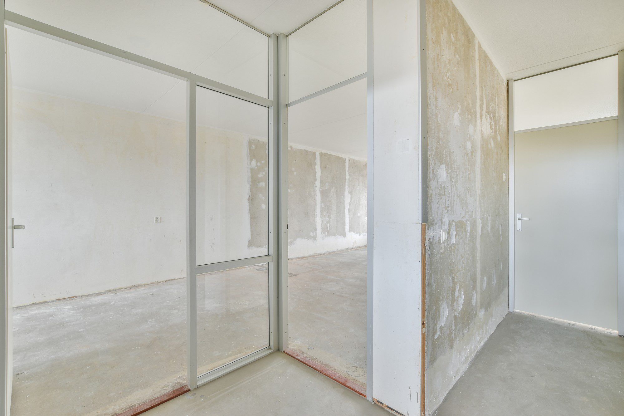 The image shows an interior space that appears to be under renovation or construction. The room has bare concrete floors and walls that are partially stripped or unfinished, revealing patches of what seems to be underlying plaster or concrete. There's a system of metal frames with glass panes forming a partition or office cubicle within the room. On the right, there is a closed door with a handle, suggesting access to another room or a closet. The ceiling is finished with what looks like acoustic ceiling tiles, and there's a diffused natural light coming in, although the source is not visible in the image. The general state of the space indicates it might be prepared for refurbishing or remodeling.