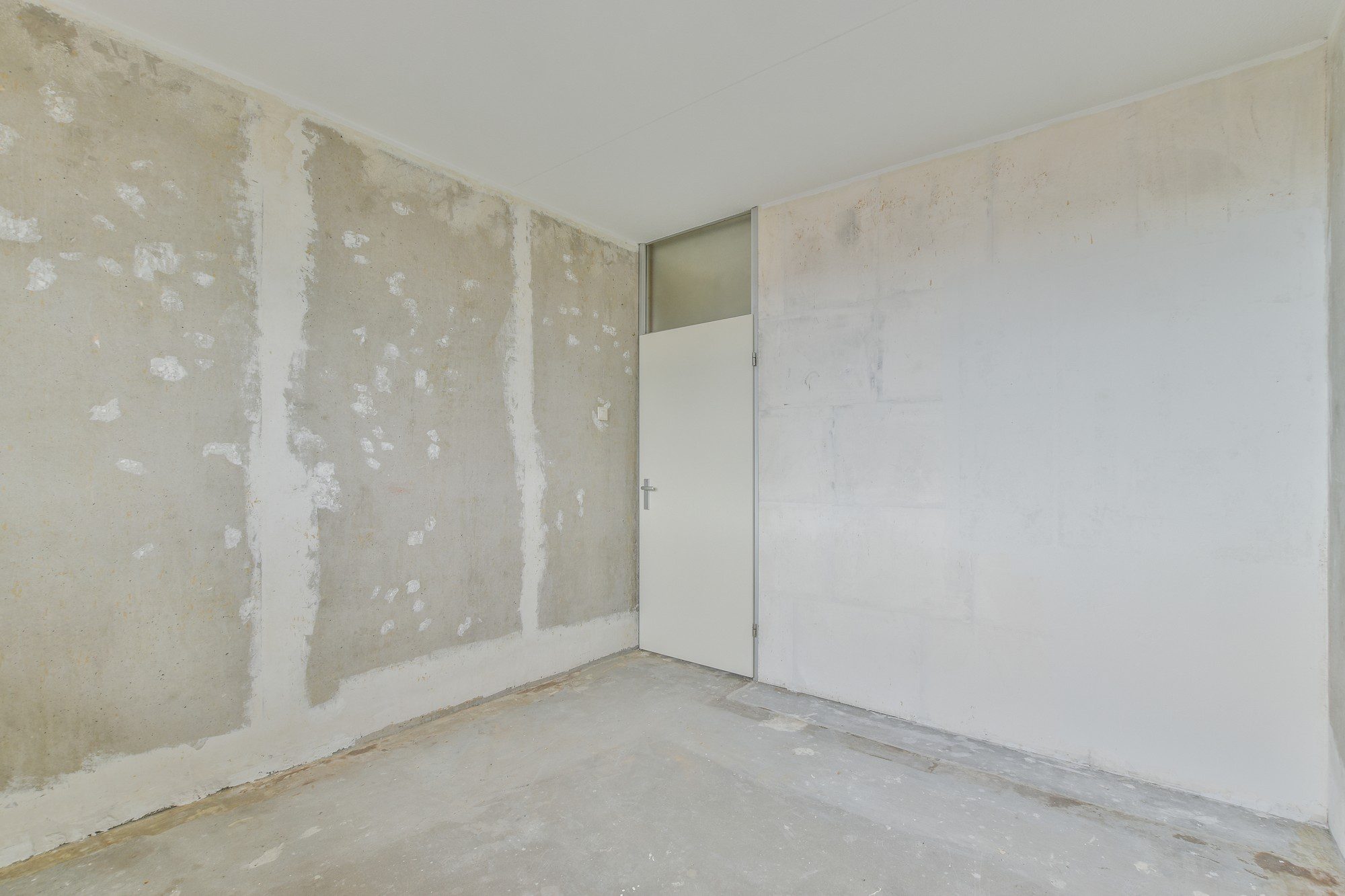 The image shows an empty room with unfinished walls. There's visible patchwork on the walls, with areas where the plaster or paint has been partially applied or removed, revealing the underlying concrete. The floor is a bare concrete surface without any covering, such as carpet or tiles. There is a closed white door on one wall, indicating an exit or entrance to another area. The ceiling appears to be finished and painted white. It seems like this room is undergoing renovation or is in need of refurbishment.