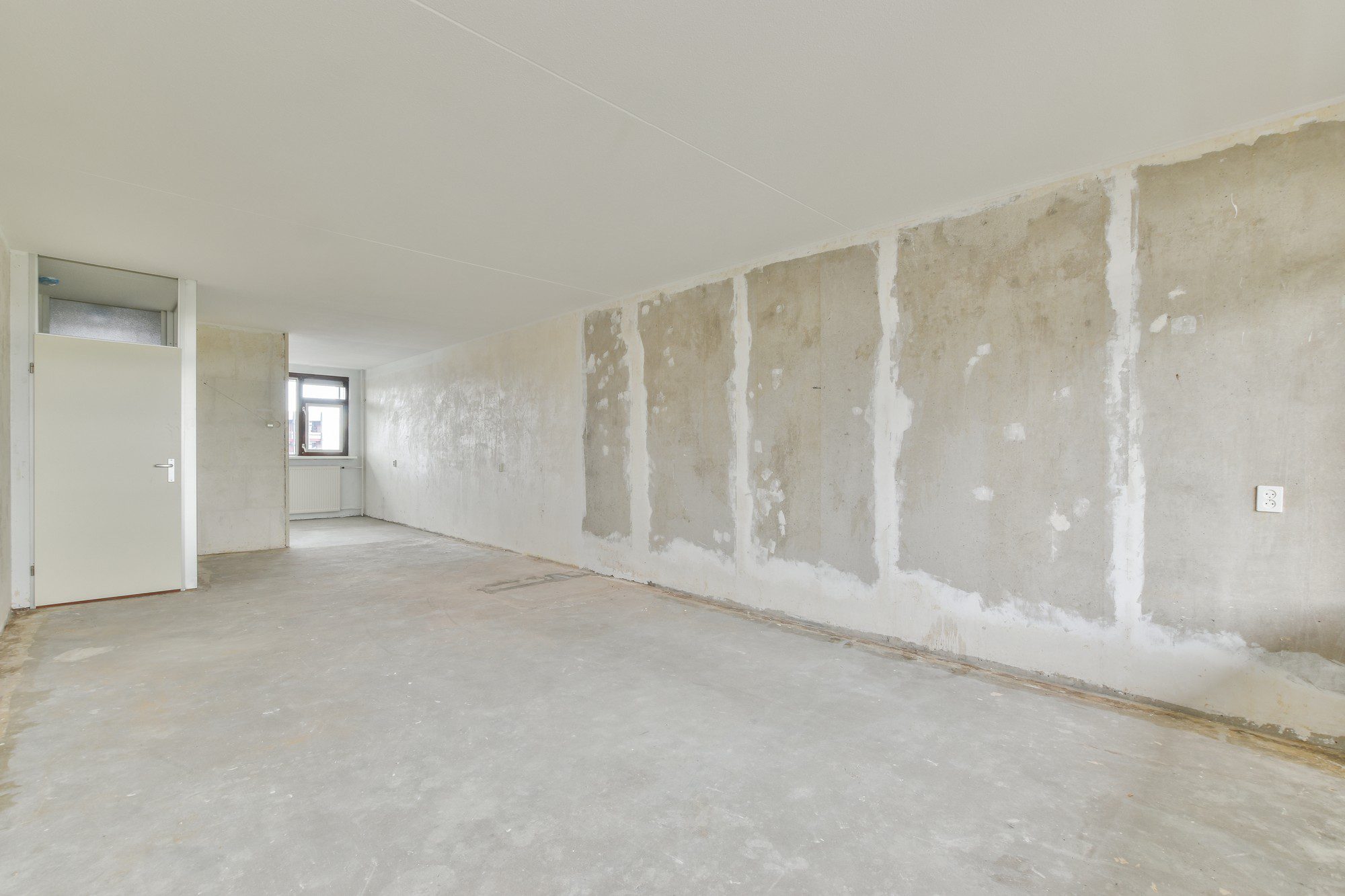 The image shows an empty room undergoing some sort of renovation or repair. The floor is bare concrete, and the walls appear to have had some plaster or paint removed, revealing patches of underlying concrete or wall material. The white patches on the walls could be where filler has been applied to smooth out imperfections before painting or wallpapering. There's a single window providing natural light, and an open doorway on the left leads to another room. Electrical outlets can be seen on the wall to the right. The ceiling looks to be in a finished state with a smooth surface.