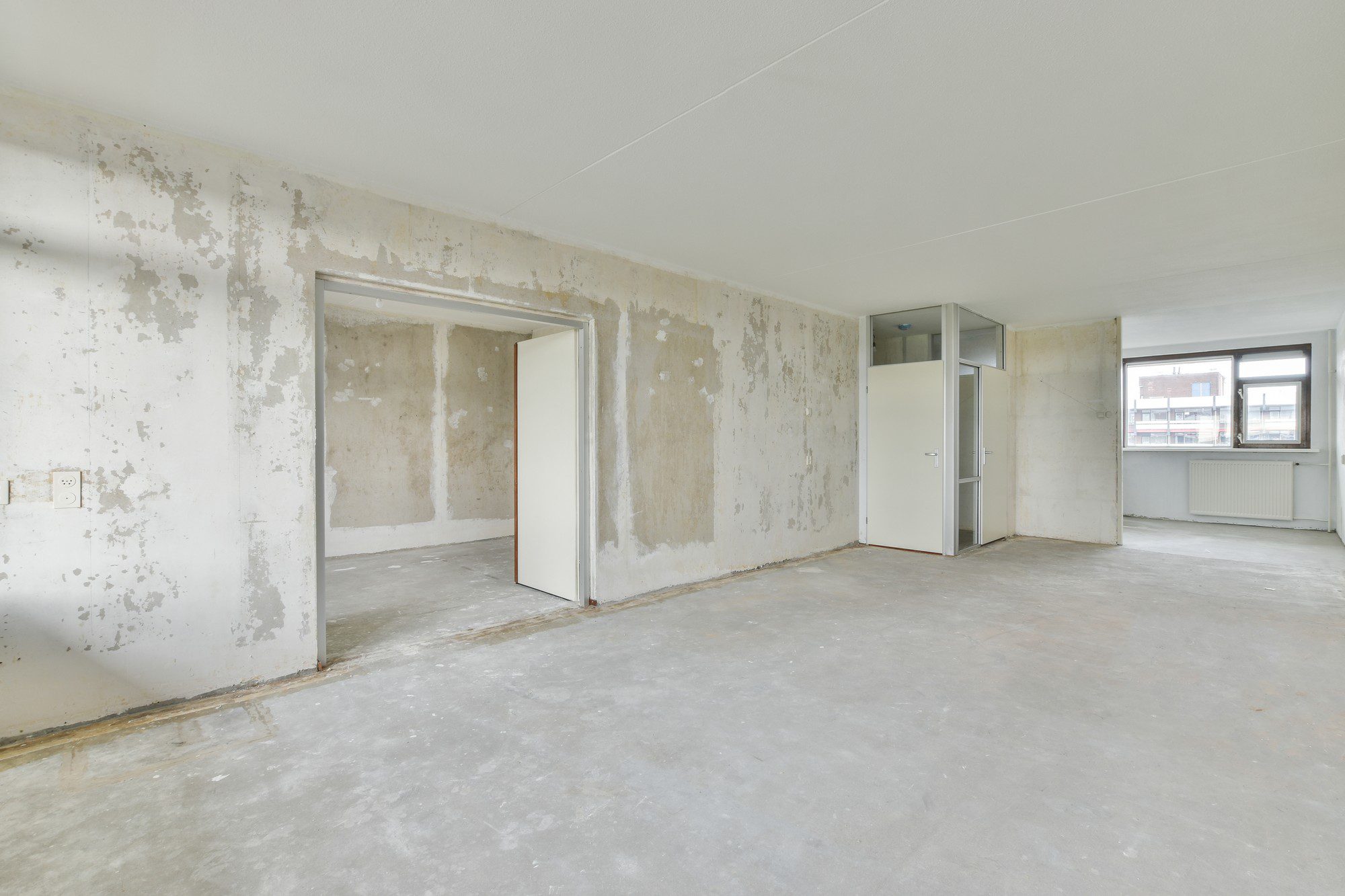 The image shows an empty room with unfinished or worn-out walls, revealing patches of the underlying wall material. There's a white ceiling with what seems to be a single light fixture. In the foreground, there's a concrete floor, and along the walls, electrical outlets and switches can be seen. There's an open doorway leading to another space, and within the room, there's a white door in a closed position, possibly leading to a closet or another section of the property. Two windows provide natural light and a view to the exterior, where other buildings are visible. The room appears to be in a state requiring renovation or finishing work.