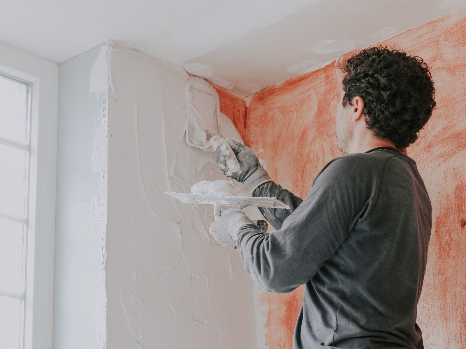 The image shows a person applying plaster or joint compound to a wall with a finishing trowel. The person is wearing gloves, which is typical for such work to protect hands from irritation caused by the materials and to ensure a better grip on the tools. The wall has been partially covered with the plaster, and you can see different shades where the material has been applied at varying thicknesses or stages of drying. The person is seemingly focused on smoothing out the compound to achieve a flat, even surface, which indicates a step in refurbishing or finishing a wall, potentially in preparation for painting or wallpapering.