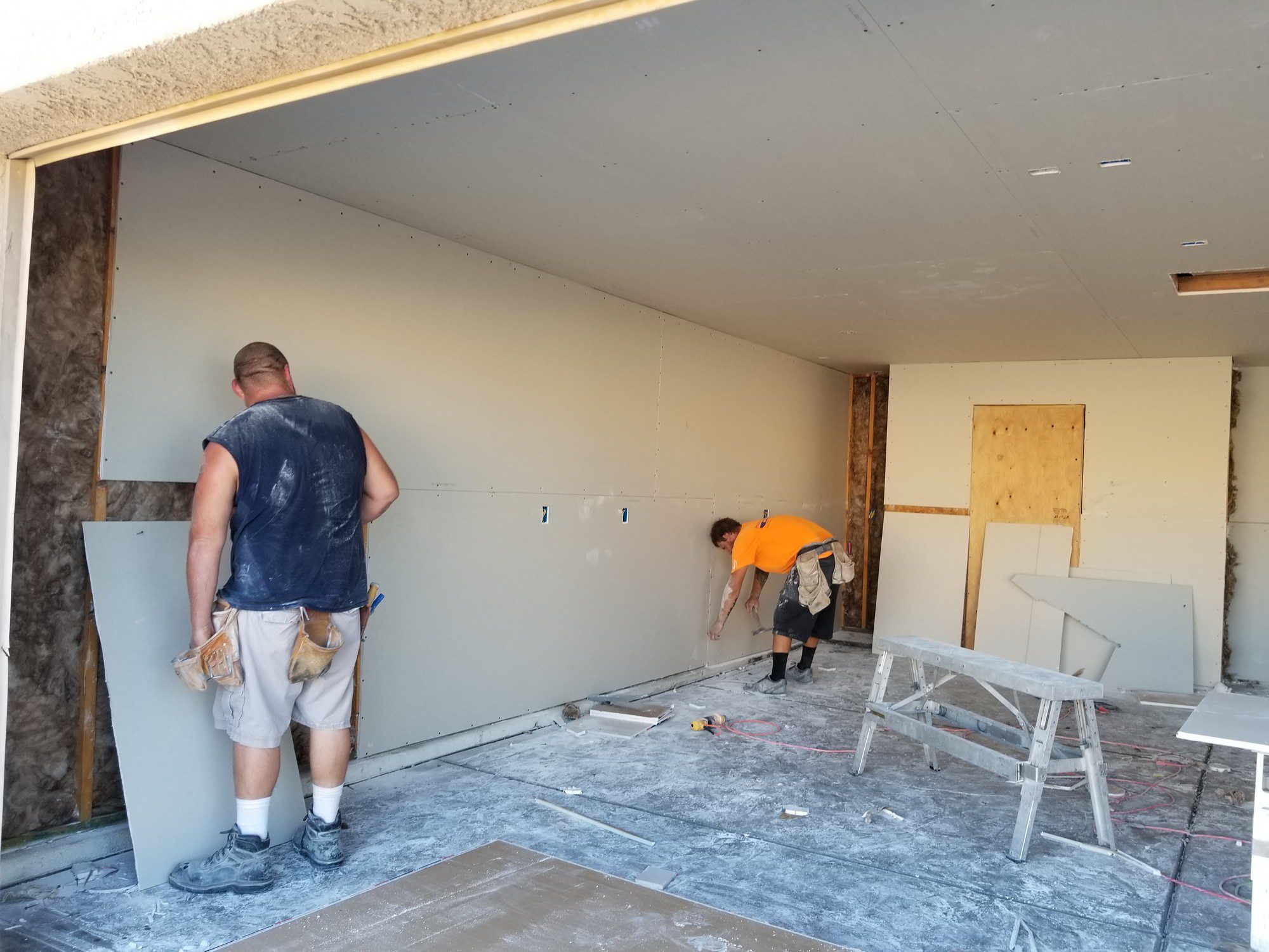 The image depicts an interior space under construction or renovation. There are two individuals who appear to be workers engaged in installing drywall or wallboard panels. One worker, wearing a blue sleeveless top, is standing and looking at a piece of drywall, while the other worker, in an orange shirt, is bending over and working near the wall.The room has exposed ceiling joists and insulation material visible in one section of the wall, indicating that the work is incomplete. Various construction tools and materials can be seen, such as a stepladder, drill, and some cut pieces of drywall. The floor is covered in dust and remnants of drywall material, typical of an ongoing construction site. The electrical outlets are partially installed, without cover plates, and electrical wires are visible. The workers are wearing casual work clothes and sturdy footwear appropriate for construction work.