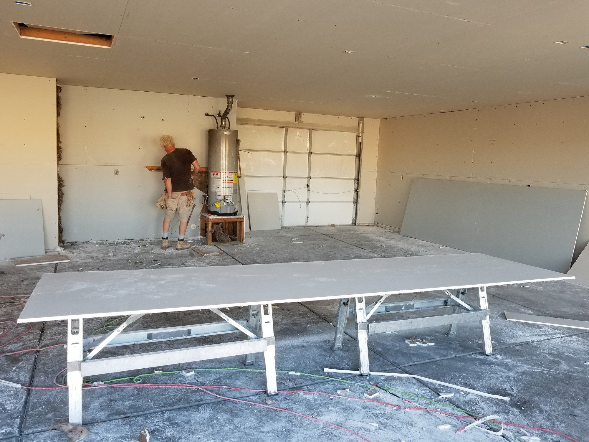 The image shows a partially constructed or renovated interior space, likely a room which is undergoing a home improvement or construction process. There's a person in the room, and they appear to be engaged in some sort of construction work or assessment. They might be measuring or cutting materials based on the tools in hand.To the left of the image, there's a large water heater appliance standing against the wall. This suggests the space could be a utility area or garage that houses major home appliances.Several sheets of drywall (gypsum board) are leaned against the wall and also laid down on a pair of sawhorses, likely to be cut to size or installed on the walls to complete the room's construction. Debris on the floor indicates recent work, like drywall or construction cuts and there are electrical cables visible on the floor, which are possibly awaiting to be properly routed or installed. The ceiling also shows exposed beams on the left side, indicating that work might not yet be completed there as well.Overall, the image depicts an active construction or remodeling scene within a building structure.