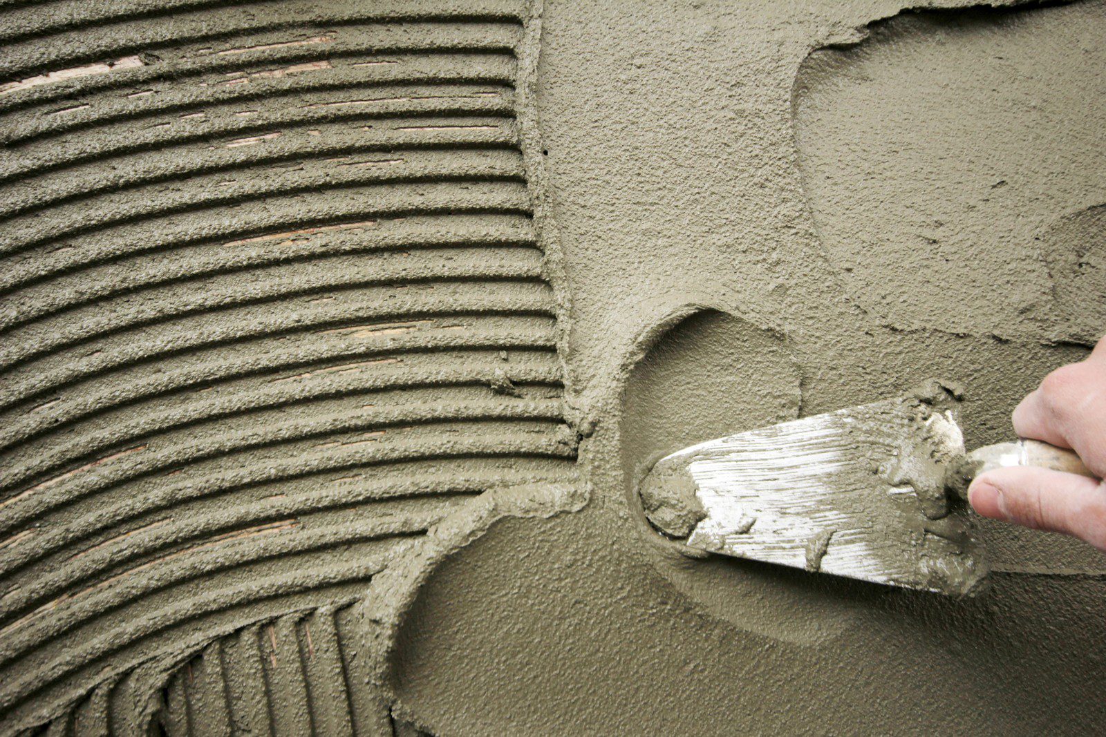 The image shows a close-up view of a person's hand using a trowel to apply and smooth out wet cement or mortar. The cement has been spread over a surface and notched with the edge of the trowel, creating parallel ridges and furrows. This technique is commonly used in masonry, tiling, and other construction settings to ensure an even adhesive distribution before placing tiles or other materials onto the surface.