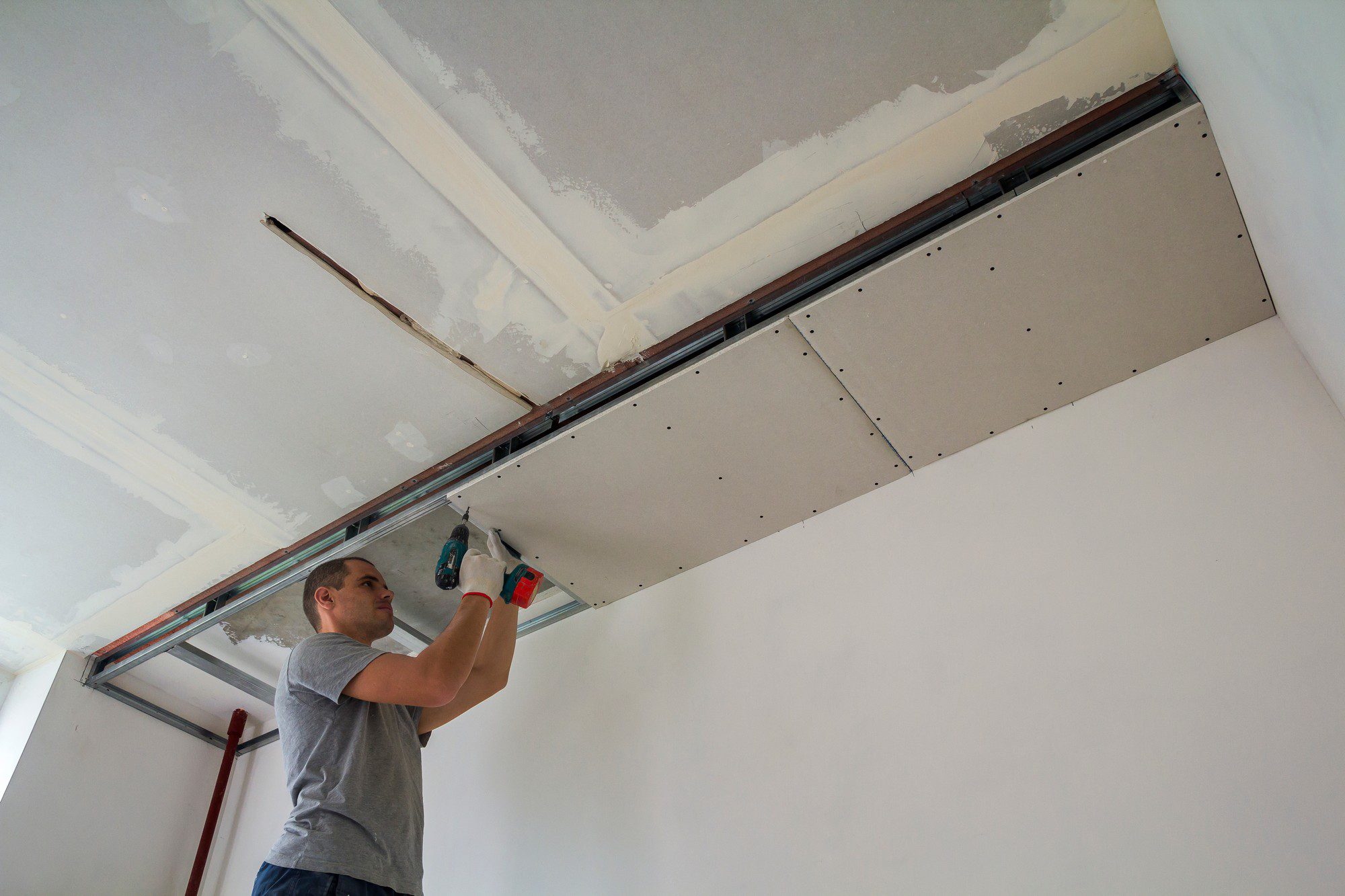 The image shows an indoor setting where a person is installing drywall, also known as plasterboard, to a ceiling. The person is using an electric drill or screwdriver to secure the drywall to the ceiling joists. You can see that sections of the ceiling drywall have already been installed, and there are visible screws and unfinished joints that will likely be taped and mudded for a smooth finish. The person is dressed casually in a gray t-shirt and is focused on their task. There is also a red pole, possibly a support pole or a tool for holding the drywall in place, within the scene. The room appears to be under renovation or construction.