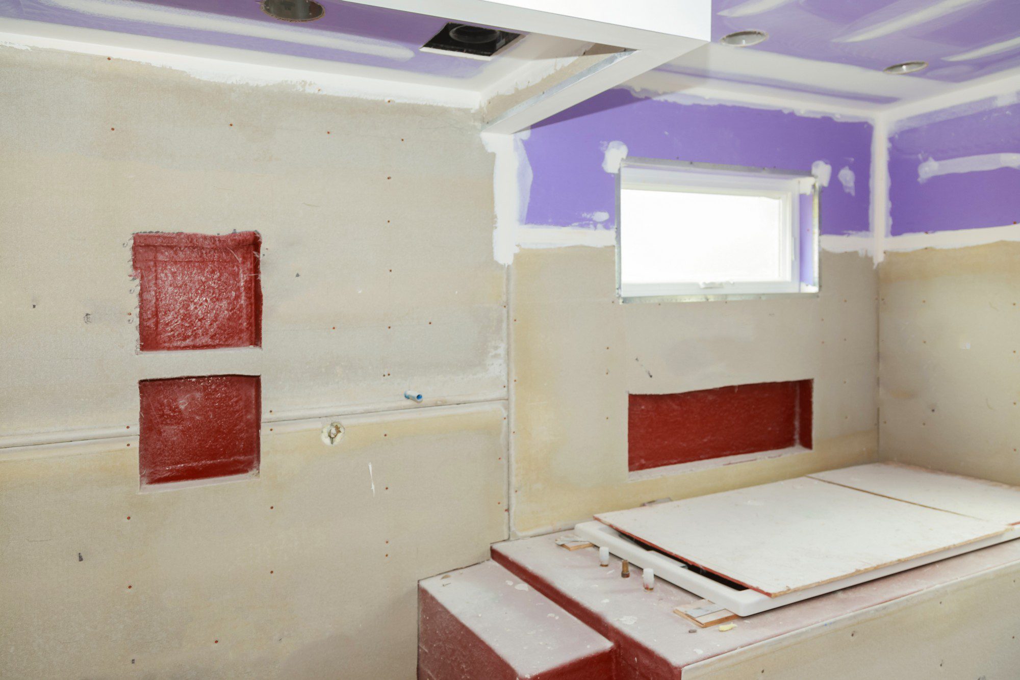 In this image, we see an interior room that appears to be under construction or renovation. The walls feature exposed drywall with some areas of purple drywall compound, likely applied for smoothing the wall surfaces before painting or further finishing. There are recessed cutouts on the wall painted in red, which might be for built-in fixtures or to conceal things like plumbing or electrical outlets. The ceiling has several downlights already installed. Additionally, there's a small window providing natural light. Construction debris or materials are also visible, like pieces of drywall, supporting the idea that this space is being worked on. The entire scene is absent of furniture or usual interior decorations, again reinforcing that the space is not yet ready for use.