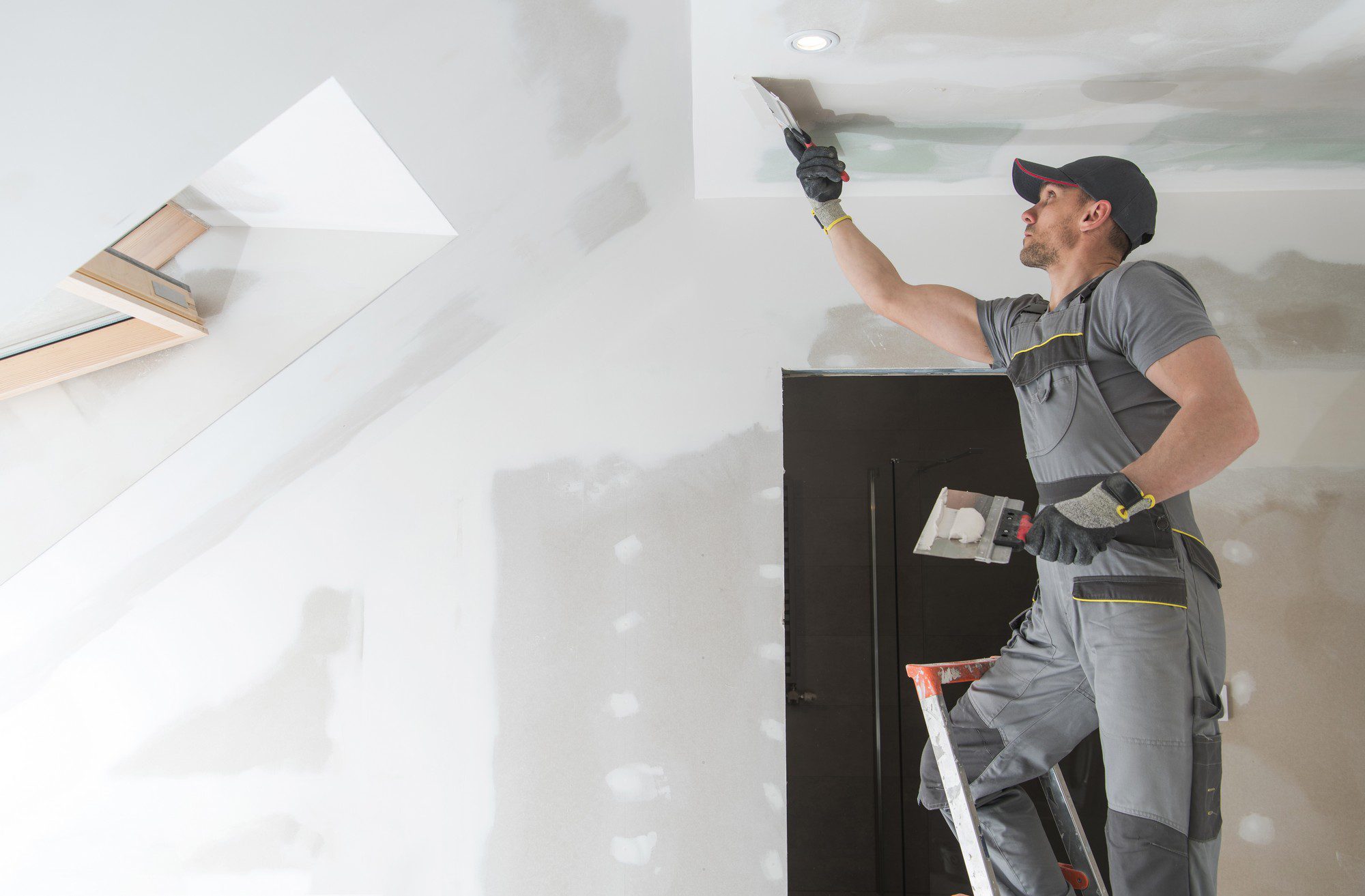 This image shows a person working on a drywall ceiling. The individual is wearing a cap, protective gloves, and a gray work outfit with yellow detailing, likely a jumpsuit or separate pants and a shirt designed for manual labour. They are standing on a step ladder and appear to be using a putty knife to apply joint compound, commonly known as spackle or mud, to smooth out the seams between sheets of drywall. In addition, we can see recessed lighting in the ceiling, and there is a skylight or an attic access panel, which suggests the room is still under construction or renovation.