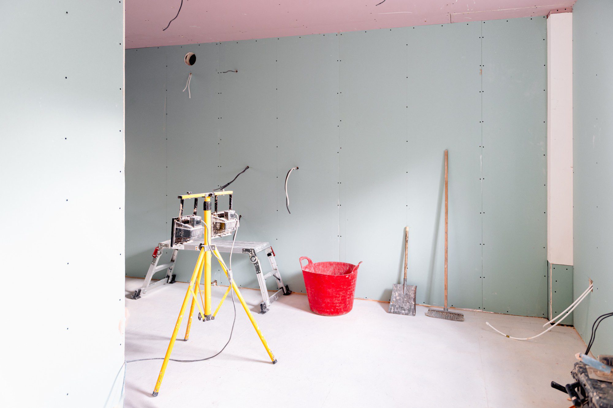 This image shows an interior space undergoing construction or renovation. The walls are partially covered with green drywall sheets, also known as gypsum board, which are secured in place but not yet finished with joint compound or paint. There is a pink ceiling which is presumably made of the same or similar material as the green walls. Visible tools and equipment include:1. A yellow and silver extendable ladder in the centre.2. A red bucket on the right side of the image.3. Construction tools, including what appears to be a shovel and a broom, are leaning against the wall to the right.4. Wiring protruding from the walls and ceiling, indicating that electrical work is part of the renovation process.5. On the left, there's a piece of construction equipment with some kind of control box; the exact function is unclear from the image, but it could be related to electrical work, painting, or another specialised task.The overall look suggests that the space is in an intermediate stage of being fitted out, with some infrastructure work underway but finishing touches like plastering, painting, and the installation of fixtures yet to be completed.