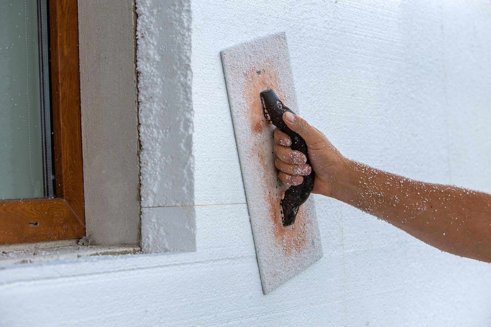 This image shows a person's hand using a tool to apply a textured coating to a wall. The wall is partially coated with the white, textured plaster, and the action of smoothing it out is causing some of the material to scatter as dust. The tool being used appears to be a specialised plastering trowel for creating textures. The context suggests that this is part of a building construction or renovation process in which the exterior wall is being finished with a textured surface.