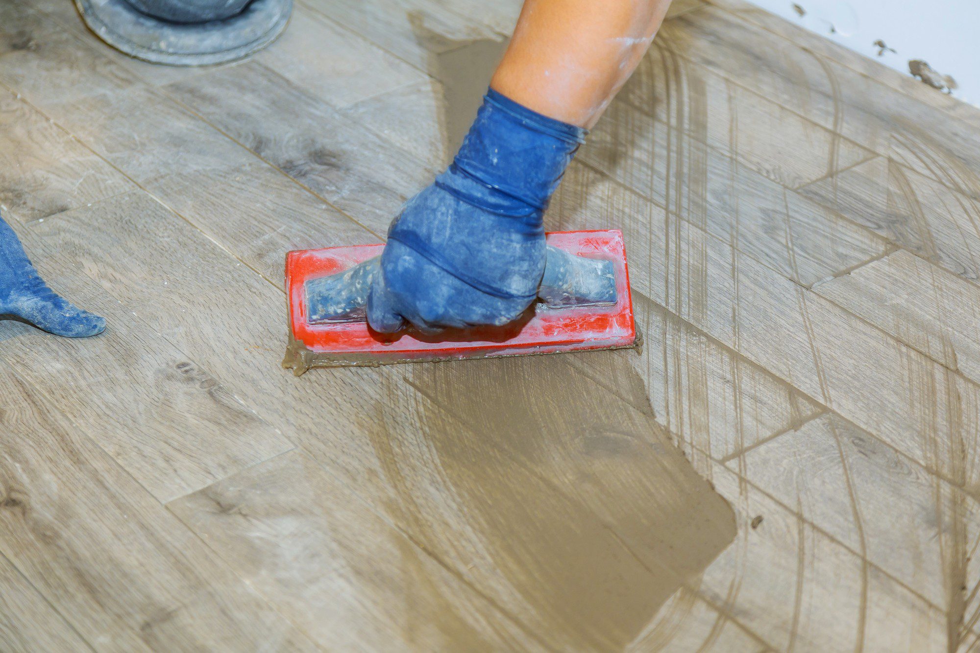 In the image provided, we see a close-up of a person's hand wearing a blue rubber glove while using a red grout float to apply or smooth grout on a tiled floor. The grout appears to be gray and is being pushed into the spaces between the tiles. The tiles have a wood-look pattern and the process seems to be part of tile installation or repair work. There is also a can or a small bucket visible at the top left corner, which might contain more grout or another related material.