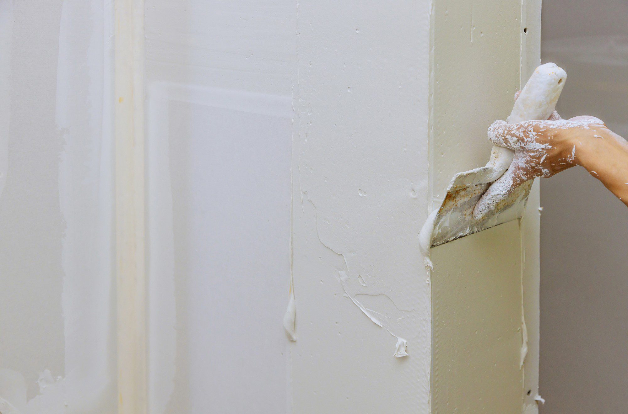 This image features a person's arm and hand using a putty knife to scrape off excess paint or material from the edge of a door or a wall. The wall and door seem to be painted white, and there are visible paint drips and excess material gathered at the edge, which the person is removing. The hand and the putty knife are covered with white residue presumably from the scraping process, suggesting some form of renovation or painting work is being done.