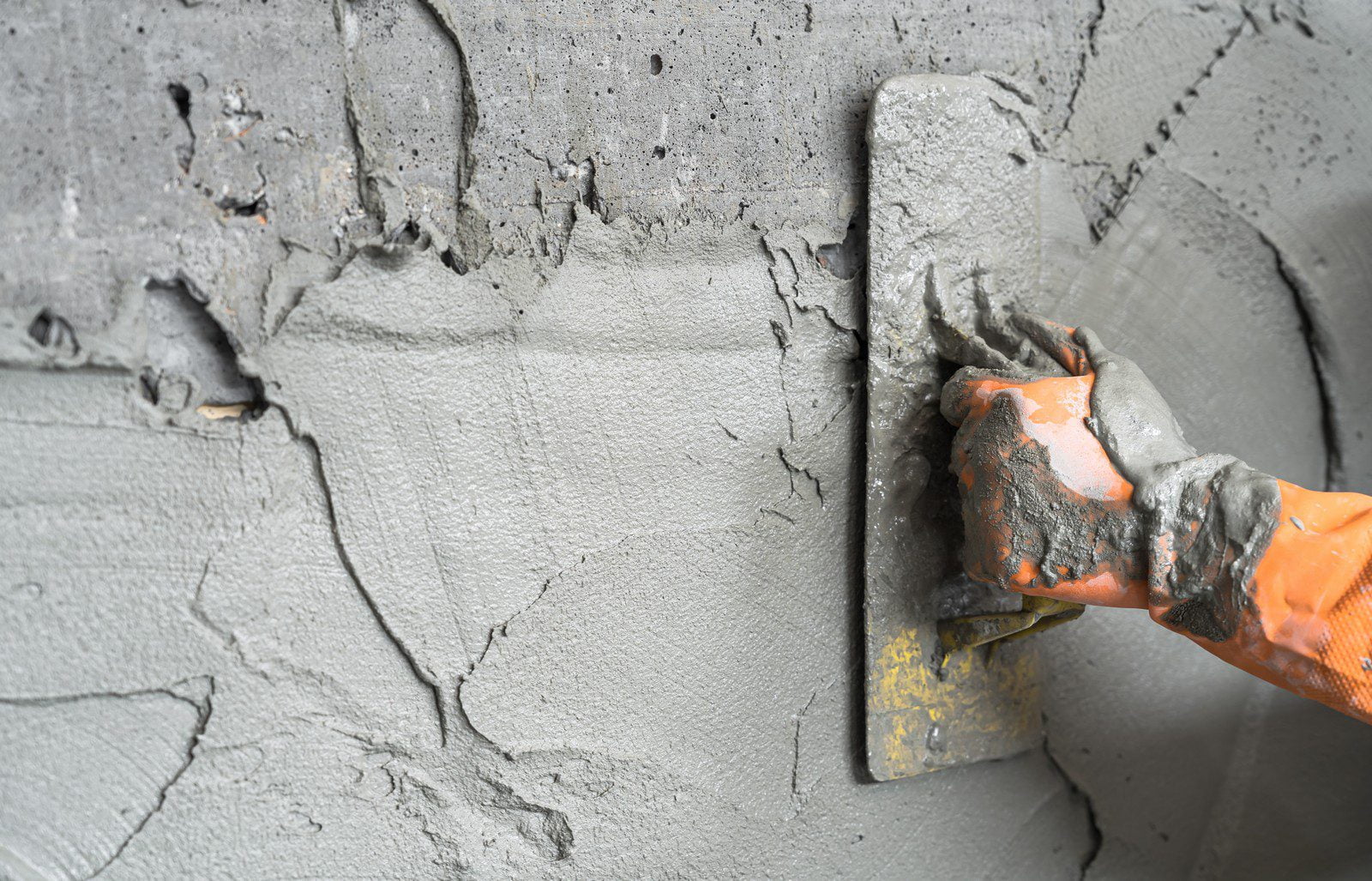 The image shows fresh cement or concrete being spread and smoothed on a surface with the use of a trowel. You can see the hand of a person wearing a brightly coloured orange glove, which is indicative of construction or masonry work, holding the trowel and applying the wet cement. The texture of the cement is rough, indicating it's still in a malleable state before setting. There are visible strokes and lines from the trowel's movement, showcasing the process of leveling and finishing the concrete surface.