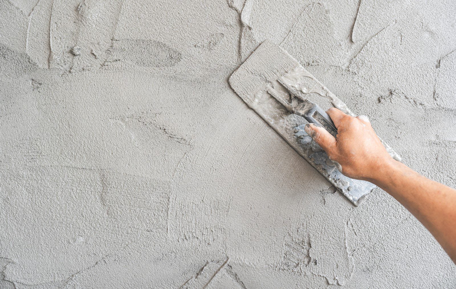 The image shows a close-up view of a worker's hand using a trowel to plaster a surface. The worker is smoothing out wet grey plaster on a wall. The trowel, which features a red handle for a secure grip, is held at an angle, indicating the plastering process is in progress. The wet plaster appears fresh and evenly spread, with a smooth texture and slightly reflective finish due to its moisture content. The background is blurred, drawing attention to the precise, detailed action of plastering. The worker's hand and the trowel become central elements, highlighting the craftsmanship and skill involved in the plastering process.