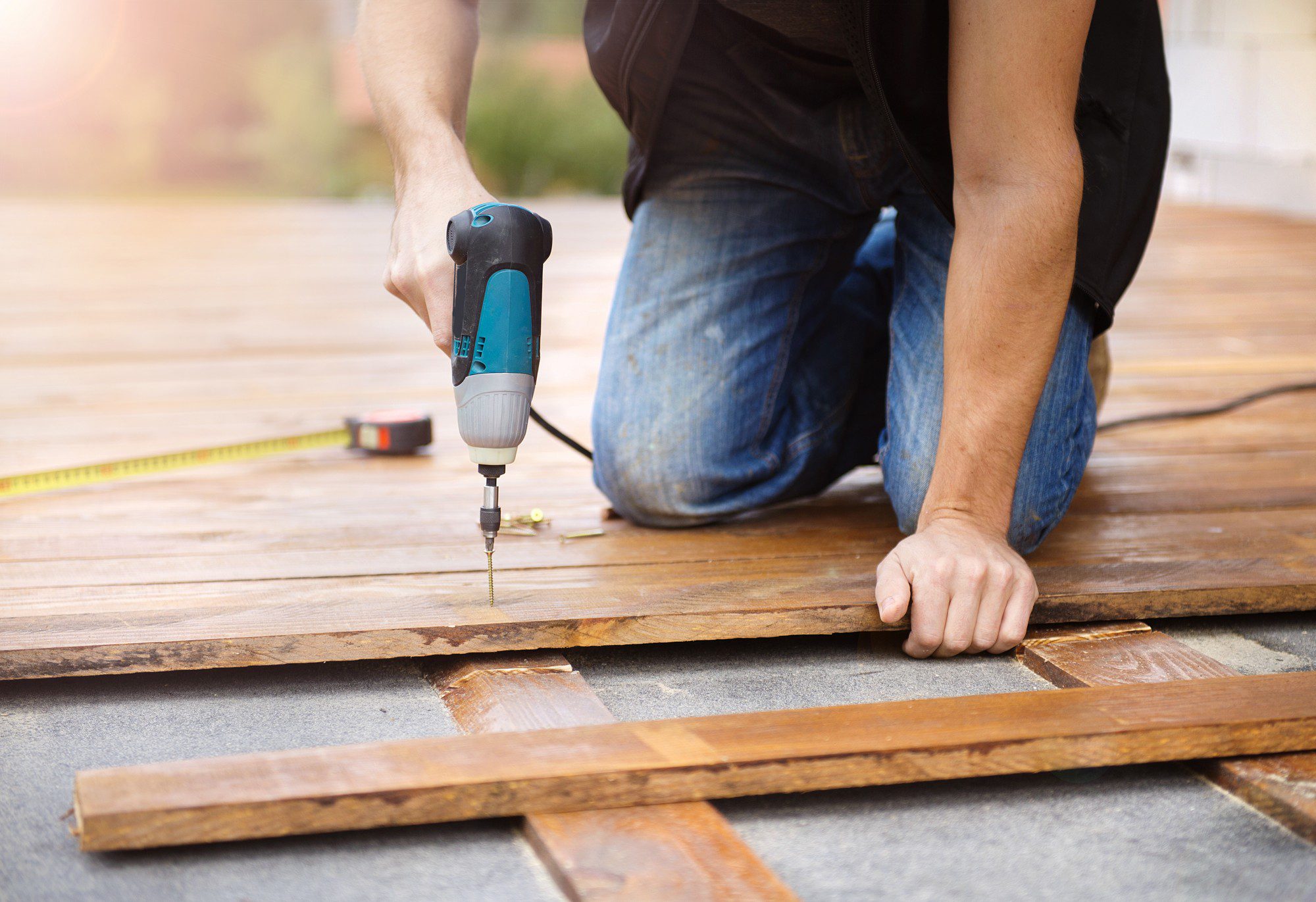 The image shows a person in the process of constructing or repairing a wooden deck or flooring. They are kneeling on the wooden planks and using a cordless drill to drive a screw into the wood. The individual is wearing casual clothes — a black top and blue jeans. In the background, there is a yellow measuring tape, partially extended, suggesting precise work is being done. The focus is on the person's hands, the drill, and the immediate area of work, with the background softly blurred, likely to emphasize the action of drilling. The setting appears to be outdoors, as indicated by the warm lighting that suggests sunlight.