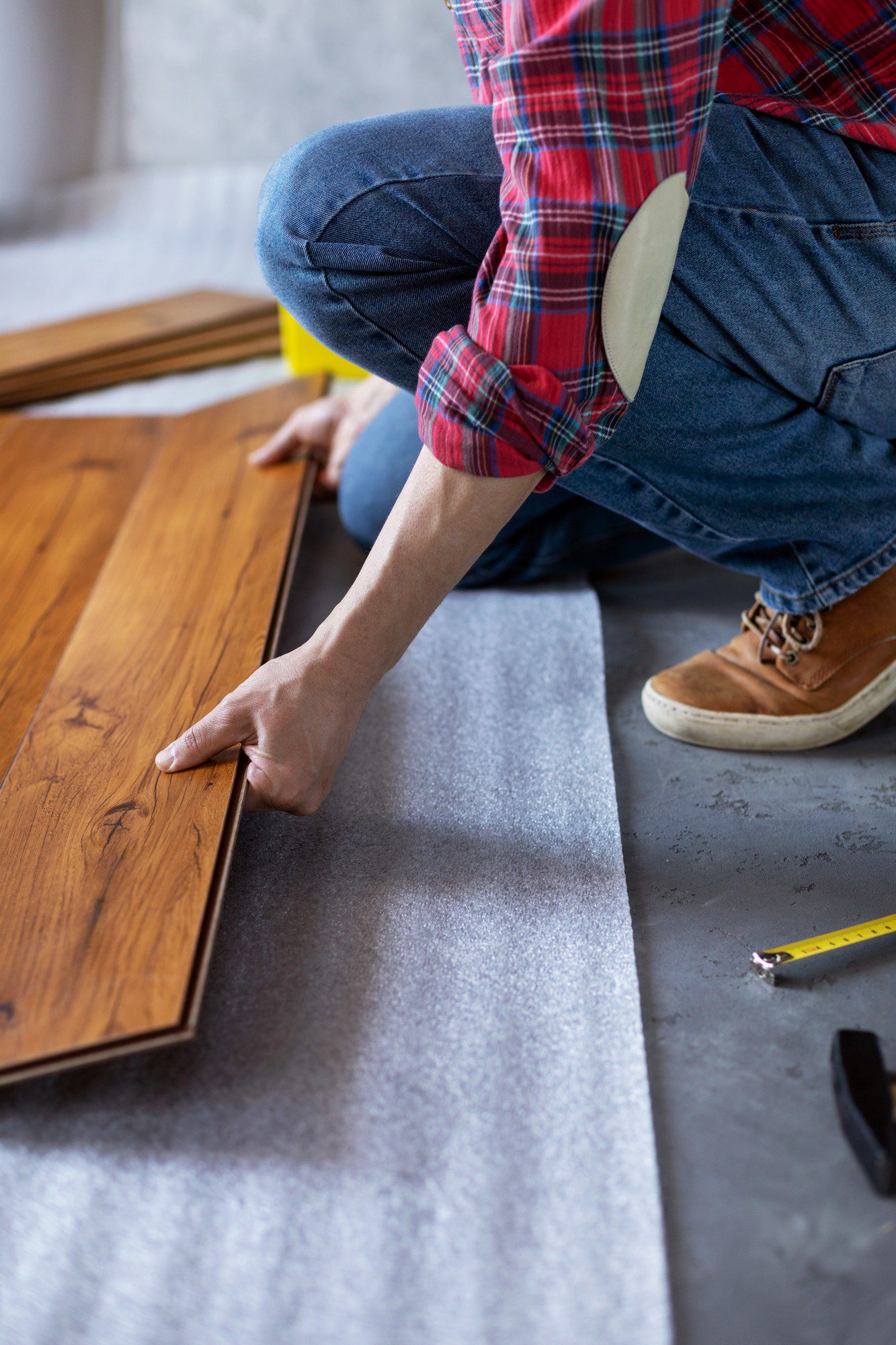 The image shows a person installing laminate flooring. The person is in a crouched position, working with a plank of laminate wood flooring, and is apparently in the process of either measuring or fitting it into place. On the floor beside them, there's a roll of underlayment, which is typically used as a base for laminate flooring to provide stability and noise reduction. Just within the frame of the image, there is also a tape measure and what appears to be a knee pad, indicating that the person is taking care to protect their knees while working on the floor. The individual is wearing a red plaid shirt, denim jeans, and lace-up boots. The setting looks like a home or building undergoing renovation or decoration.