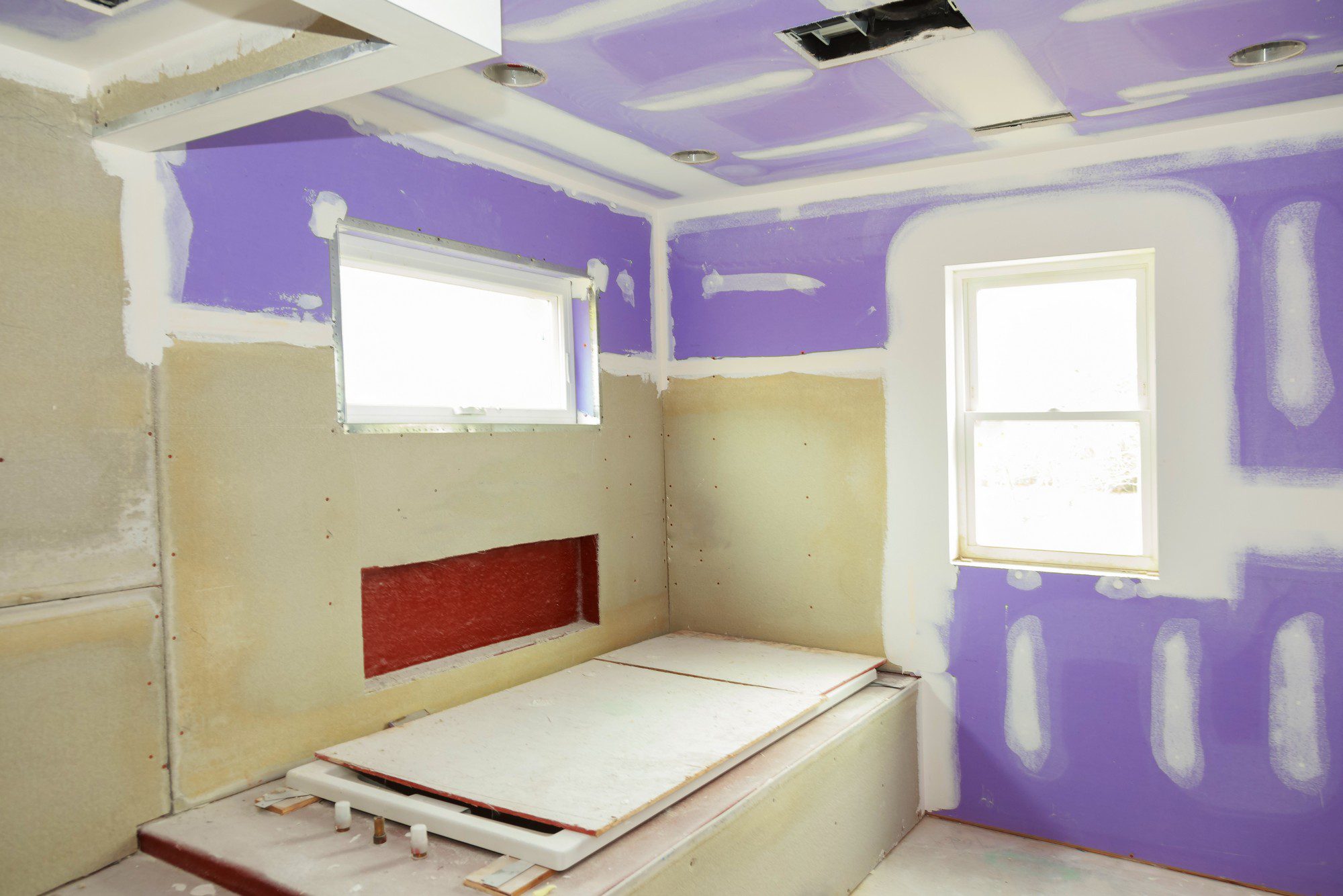 The image shows a room under construction or renovation. The walls are insulated with fiberglass batts, partially covered with purple-coloured drywall compound (widely known as drywall mud) which seems to be applied for the purposes of sealing joints and screw indentations before painting. Some of the wall studs and insulation materials are still exposed. There are two windows allowing natural light into the room, and several recessed lighting fixtures can be seen in the ceiling. The floor is covered with protective material, possibly to keep it clean during the work. The room looks unfinished and is likely in the process of being remodeled or completed.