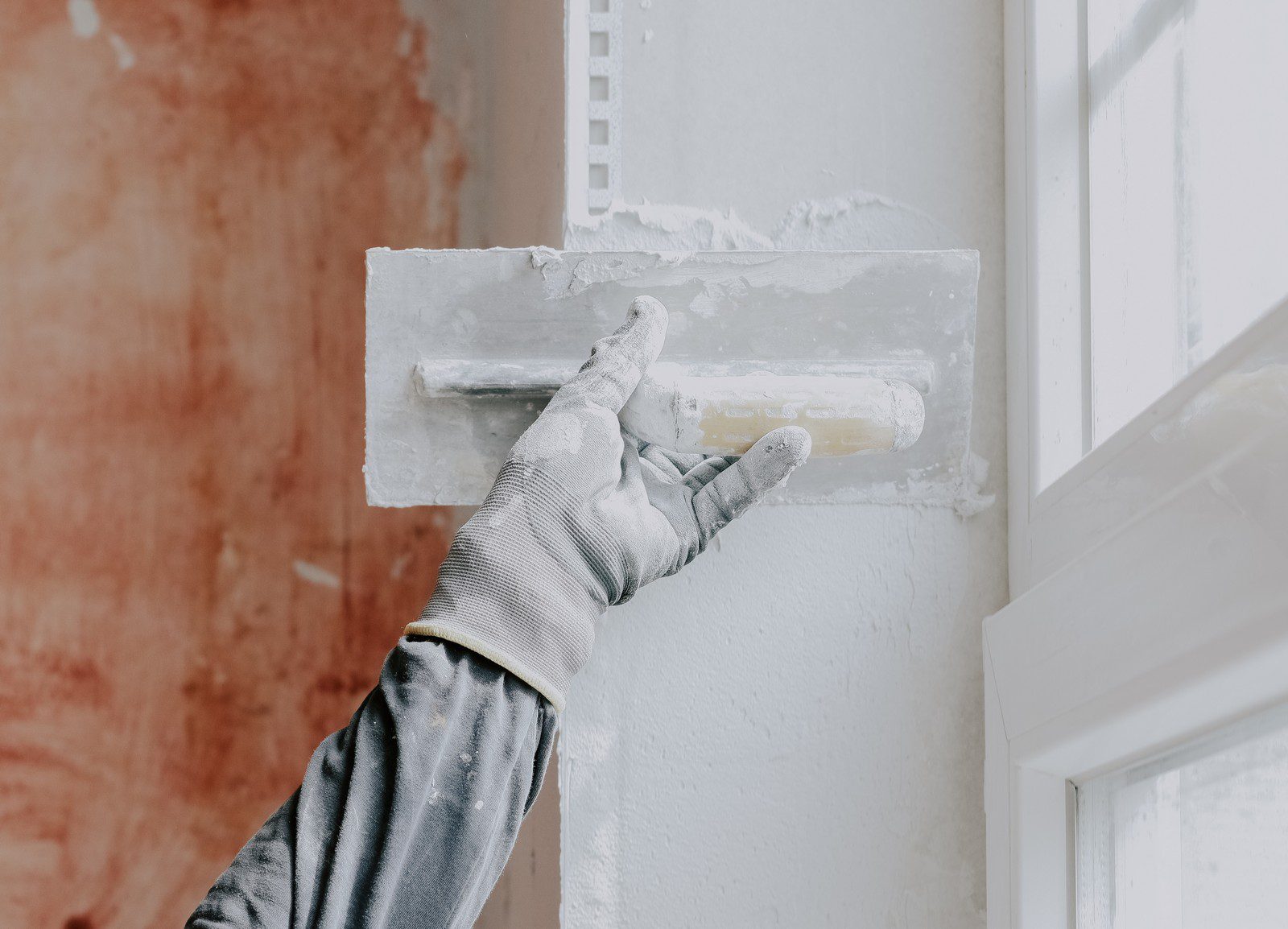 The image features a close-up of a plasterer's hand wearing a construction glove. The glove is well-worn, indicating extensive use. The hand is holding a trowel covered in wet plaster, which is being applied to a wall. The wall surface is partially covered with fresh plaster, and the texture of the wet plaster can be seen clearly, appearing smooth and slightly reflective. In the background, there are indistinct, blurred elements typical of a construction or renovation site, such as tools, scaffolding, and unfinished surfaces. The overall scene captures the meticulous and skilful nature of plastering work.