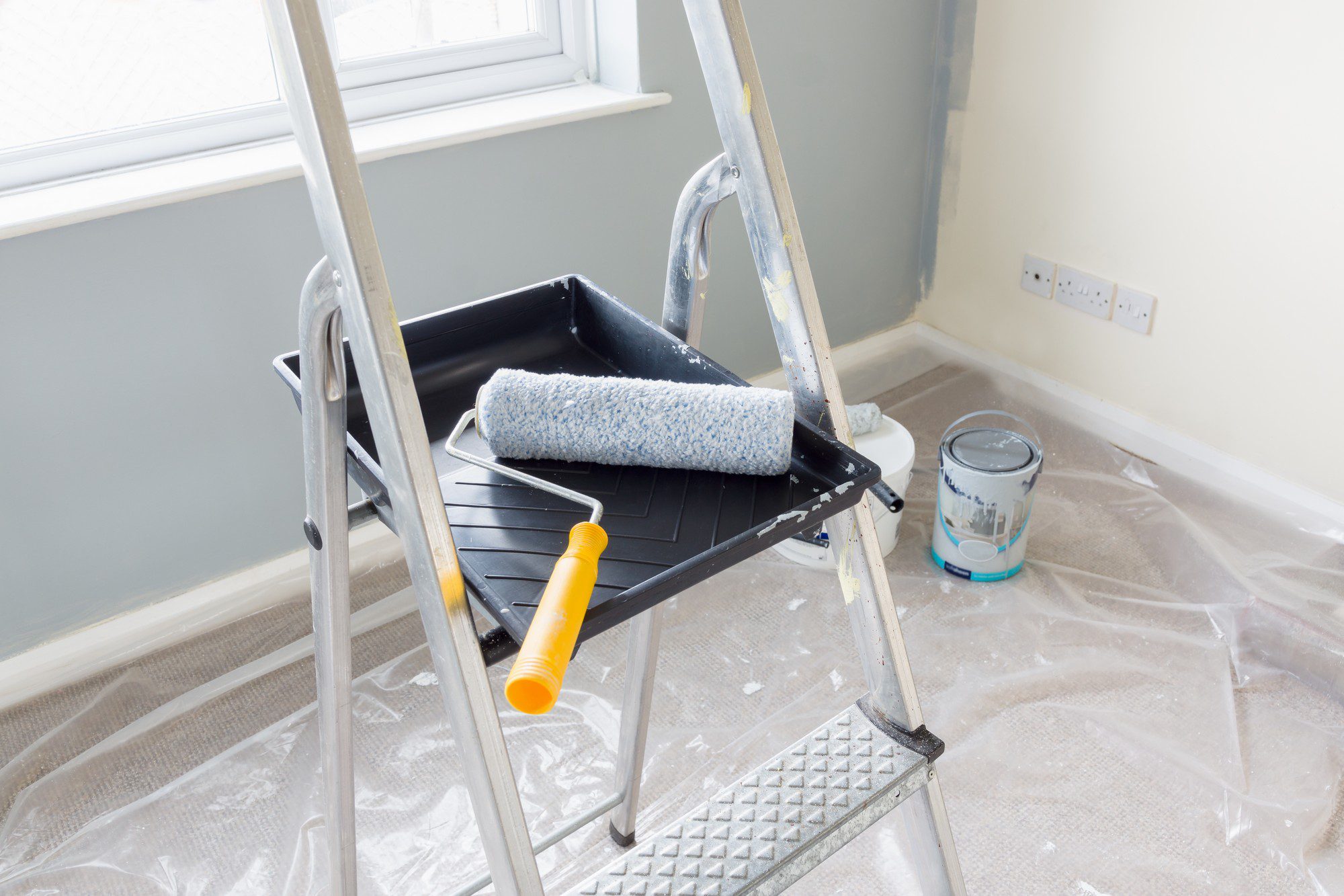 The image shows an indoor painting setup, including a step ladder with a paint tray and roller atop it. The floor is protected with a plastic drop cloth, and there's an open can of paint on the floor, indicating that someone is in the process of painting or renovating the room. The wall in the background appears to be freshly painted with a light-coloured paint. There are also electrical outlets on the wall and a section of the wall that has not yet been painted, as the edge is taped off for a clean line.