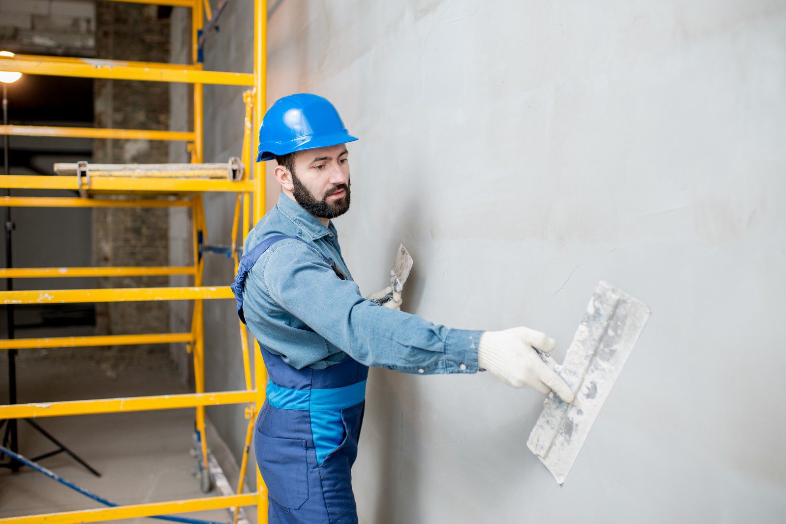 The image shows a man, presumably a construction worker or plasterer, wearing a blue hard hat and workwear including gloves. He is applying plaster on a wall using a trowel. In the background, there is a yellow scaffolding structure, indicative of ongoing construction or renovation work. The man appears focused on his task, ensuring a smooth application of the plaster. The scene suggests an interior workspace that is in the process of being finished or remodeled.