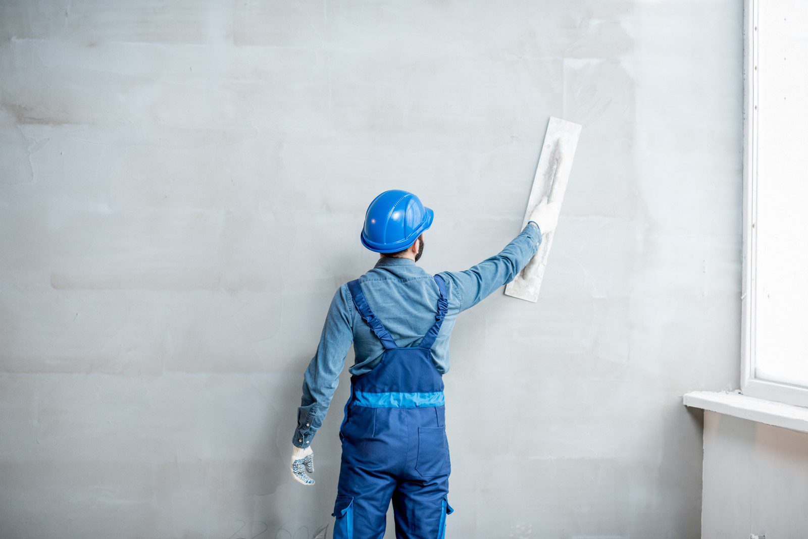 This image shows a person from the back who appears to be engaged in some sort of manual labour, likely related to construction or refurbishing. The individual is wearing a blue hard hat, which suggests a concern for safety often associated with construction sites. The person is also wearing a blue coverall with reflective elements and is holding what looks like a spatula or plastering trowel, which indicates they might be in the process of applying plaster or smoothing out a wall surface. The wall itself appears partially finished or in the process of being smoothed. To the right, there is a window, and at the bottom right corner, there seems to be some sort of white construction material, possibly more plaster or construction-related supplies.