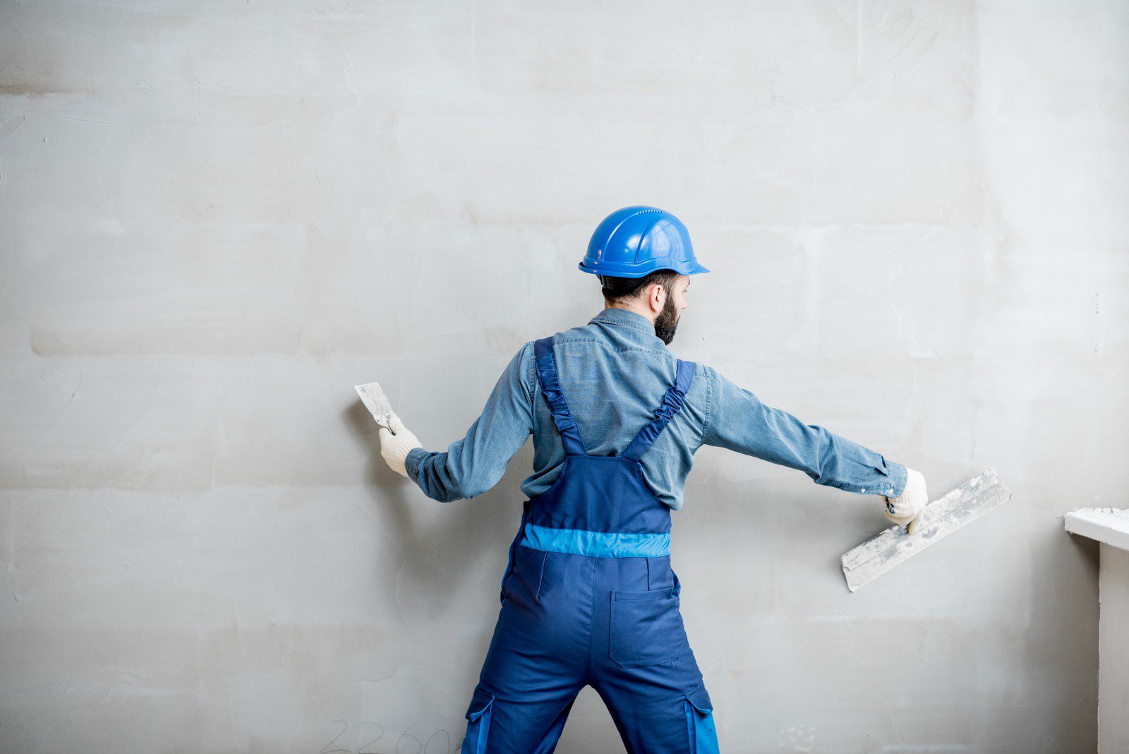 This image shows a person working on a wall. The person is dressed in a blue work uniform, which consists of a long-sleeve shirt and pants, with a blue hard hat on their head, indicating they could be a construction worker or a tradesperson. They are using a plastering trowel in each hand to apply or smooth plaster or a similar material on the wall. The wall looks to be in the process of being finished or repaired. The worker's stance suggests they are in motion, actively spreading the material on the surface of the wall.