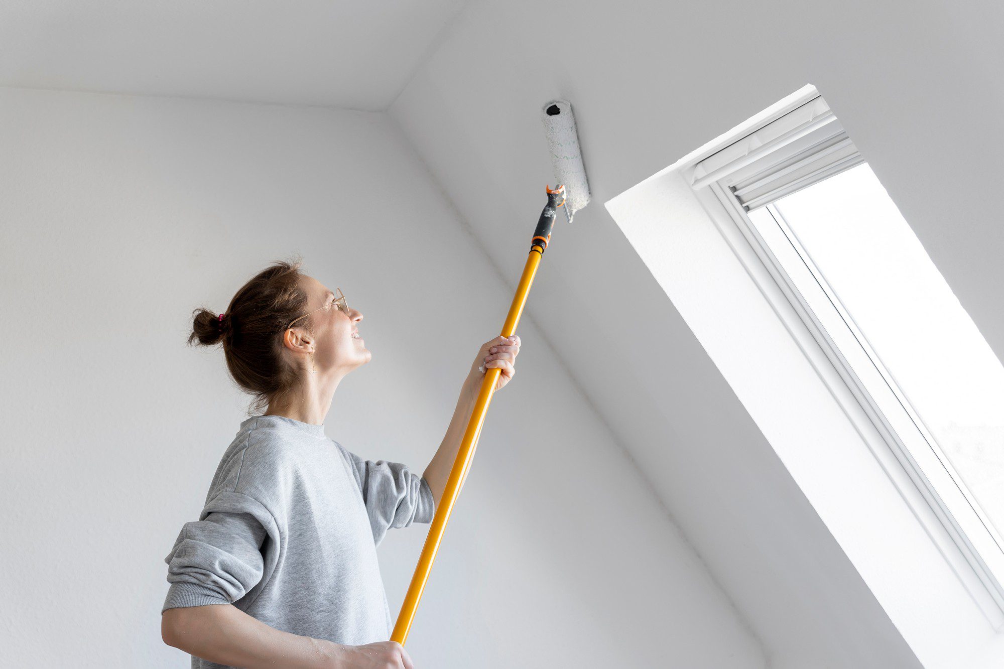 The image shows a person painting a ceiling with a roller brush. The person appears to be a woman and she is using a long yellow handle on the roller brush to extend her reach. She is wearing casual clothing—a grey long-sleeved shirt—and glasses, and her hair is tied back in a bun, indicating that she is in the middle of a home improvement task. The setting looks like an interior room with white walls and the ceiling is also white, highlighting that the painting is likely for maintenance or a freshening up of the space. There is a large, vertically oriented window with white trim in the background, allowing natural light into the room.