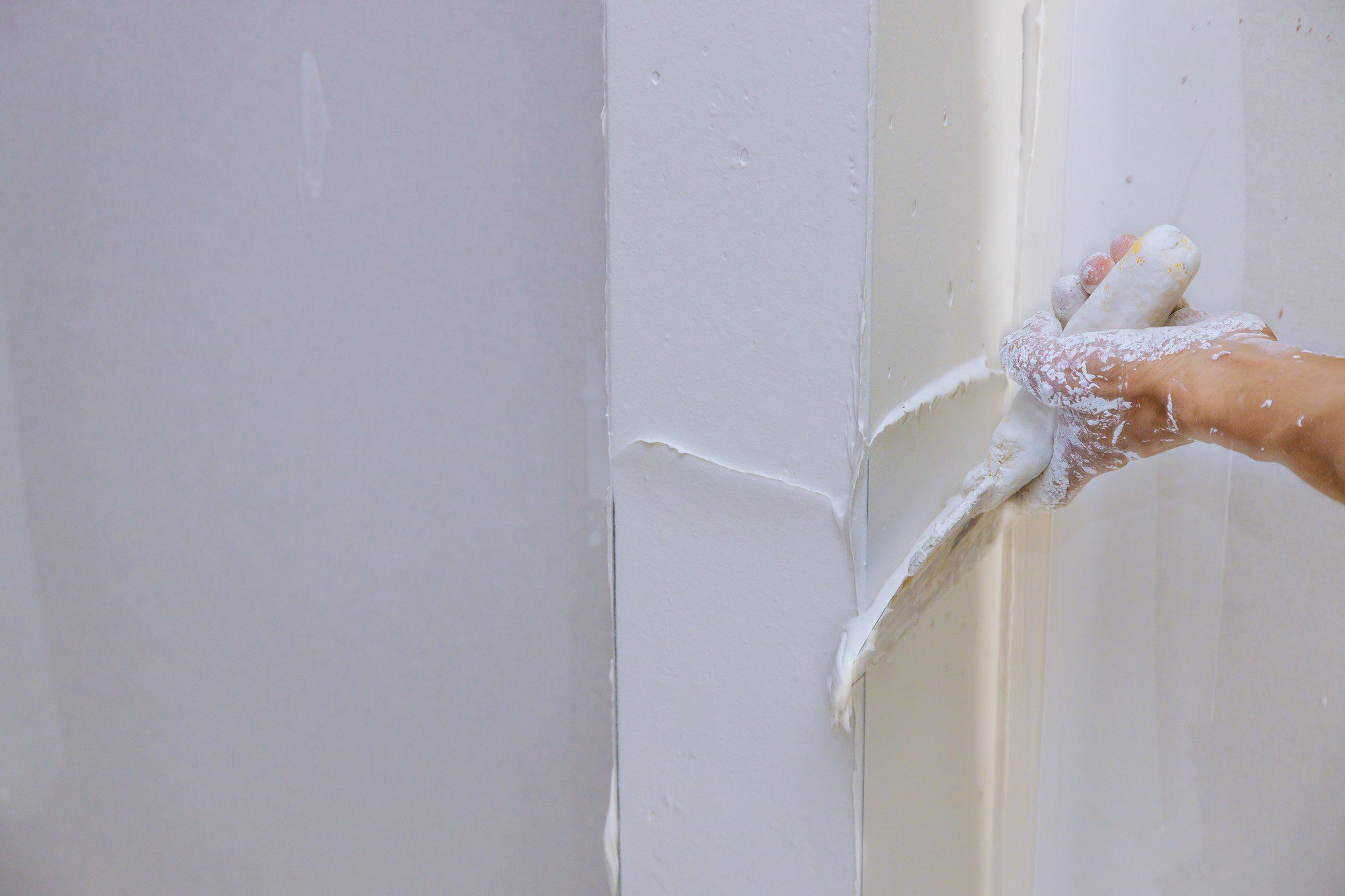 The image shows a close-up view of someone working with joint compound or spackling paste, commonly used in drywall installation and repair. The person's hand is wearing a glove and is using a taping knife to smooth the compound into the corner of a wall where two sheets of drywall meet. The white paste is typically used to fill seams and cover up nail or screw indentations. There are some excesses and splatters of the compound on the glove and wall, which is common during the application process before the compound is smoothed and later sanded. The corner appears to be reinforced with paper or mesh drywall tape beneath the compound, which helps prevent cracking and provides strength to the joint.