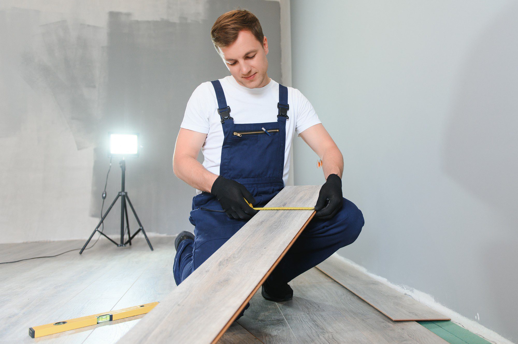 **Alt Text:**A repairman is seen laying laminate flooring in a home. He is crouched down on one knee, focused on fitting a plank into place. The man is wearing a light blue t-shirt, grey work trousers with black knee pads, and sturdy black work boots. He has protective gloves on his hands and appears to be in his mid-30s with short brown hair. The room is well-lit with natural light streaming in from a large window on the left side, casting a soft glow across the wooden floor. Various tools are scattered around him, including a tape measure, a pencil, and a hammer. There are several neatly stacked boxes of laminate planks nearby, indicating that the job is still in progress. The walls of the room are painted a light grey, and in the background, there is a glimpse of a white door standing ajar.