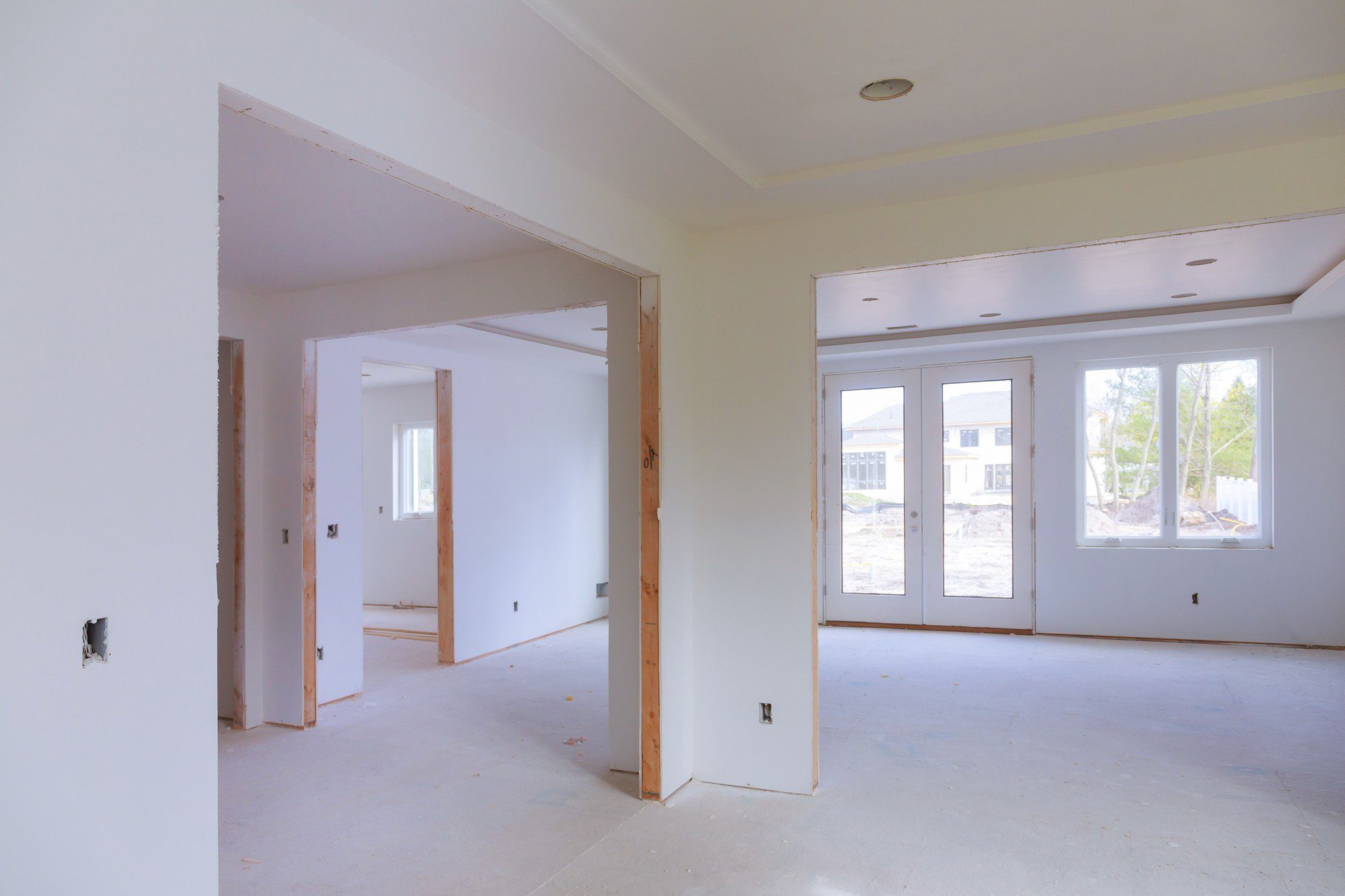 The image shows the interior of a room under construction or renovation. The walls are unfinished, with visible drywall and joint compound but no paint or wallpaper. There are cut-out holes for electrical outlets. We can also see a support column and a header, indicating a doorway or passageway to another room. The flooring appears to be subflooring without any final floor covering such as carpet, tile, or hardwood. There is a dual-pane sliding glass door with a view of the exterior area, where some construction or excavation appears to be ongoing, and other houses are visible in the background. The ceiling has a recessed lighting fixture installed. This scene is typical of a house that is not yet completed and is likely in the process of being built or remodeled.