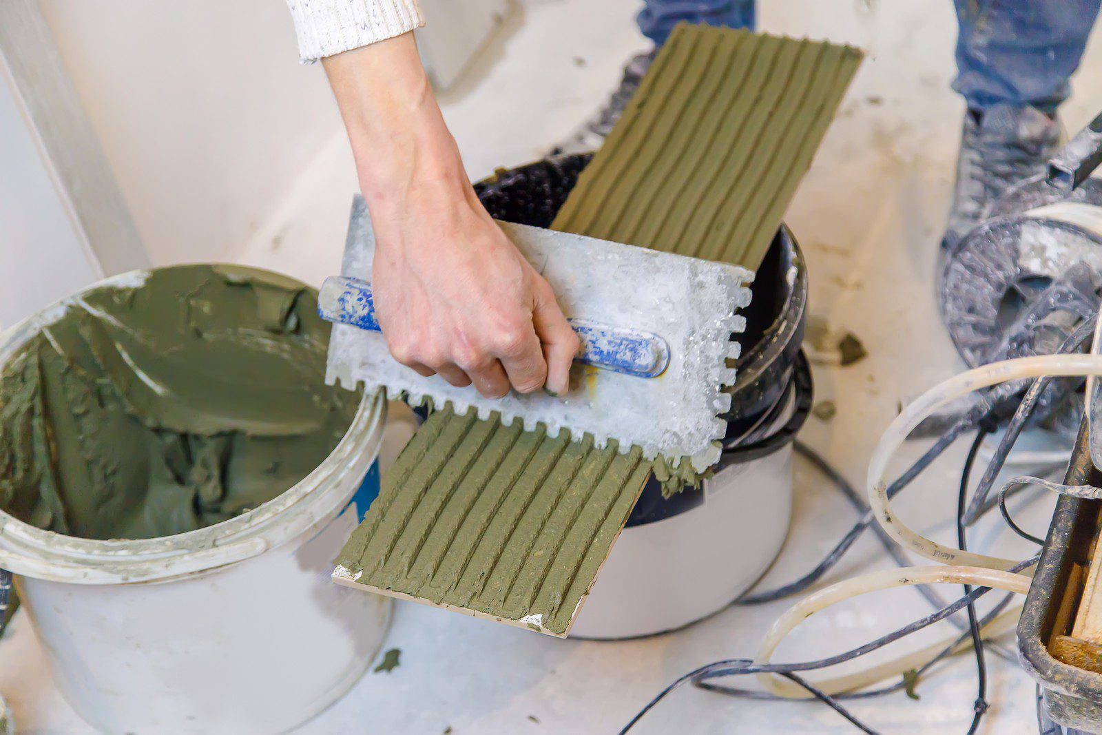 The image depicts a person engaged in construction or tiling work. They are holding a notched trowel with grey construction adhesive or mortar applied to it, creating ridges that will help tiles adhere to a surface. A bucket with more of the adhesive or mortar is also visible, along with a mixing tool or electric mixer with a spiral mixing attachment lying on the floor, coated with the same material. There are flecks and smears of the adhesive around, indicating an ongoing work process. The individual is likely preparing to apply or has been applying this material to a surface or to tiles being installed.