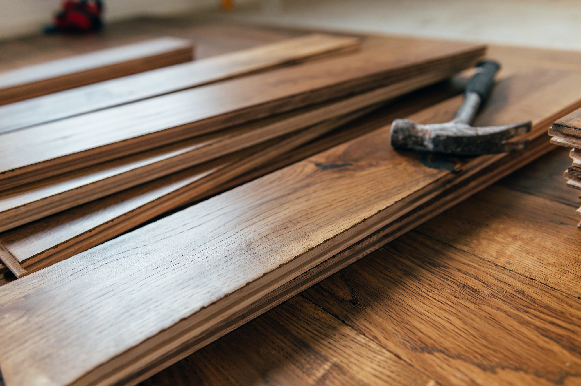 The image shows a stack of wooden laminate flooring planks laid out on a wooden floor, with a hammer resting on the planks to the right. There are also more flooring materials or tools in the background, but they are out of focus. The setting suggests that someone might be in the process of installing or renovating a hardwood floor.