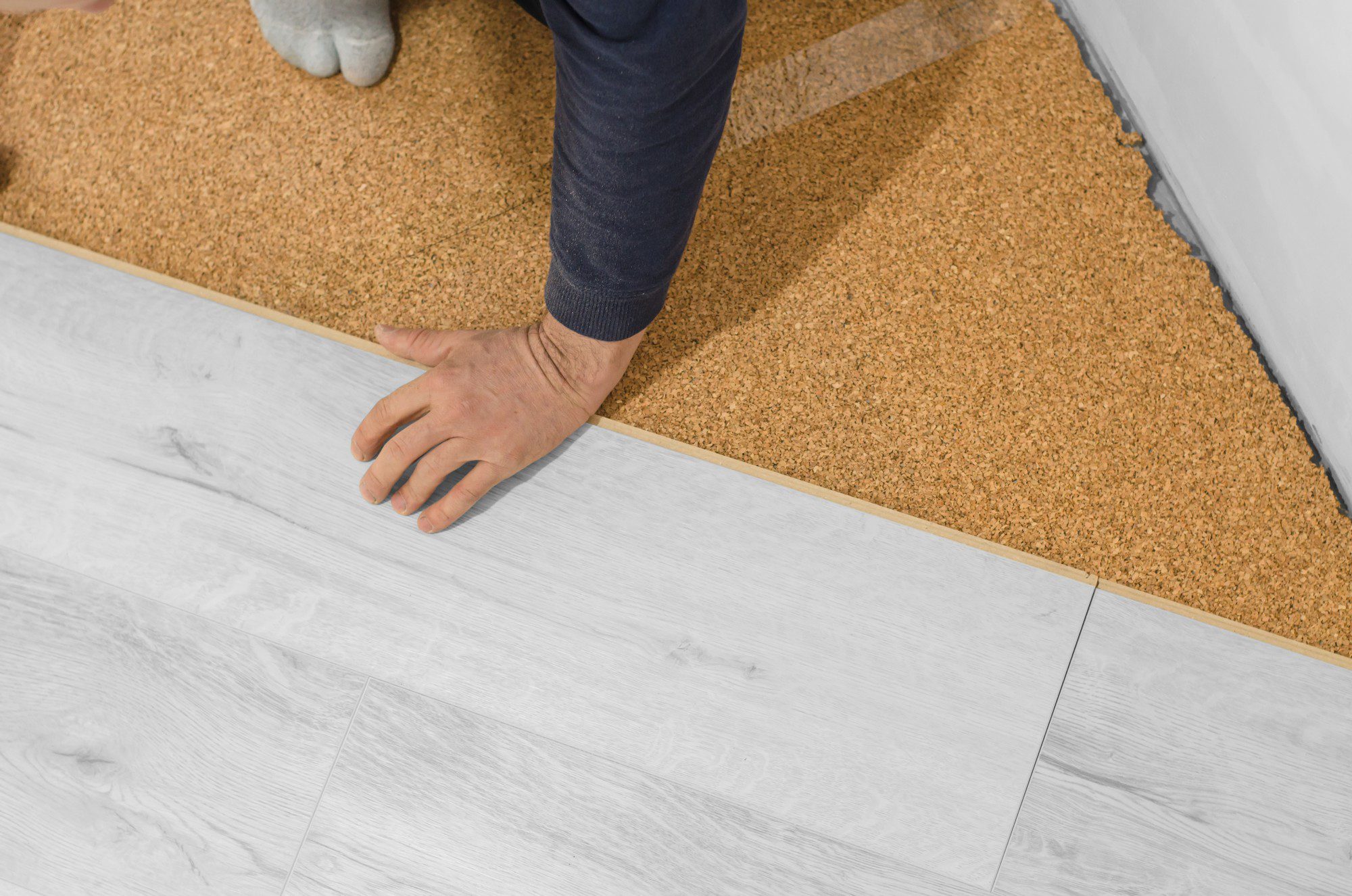 The image shows a close-up view of a floor at the threshold between two rooms or areas. On one side, there's a light-coloured laminate or vinyl plank flooring, and on the other side is cork flooring. A person's arms and legs are visible as they appear to be installing or laying down the cork flooring. There's also a glimpse of a door frame on the right side of the image, indicating this could be an entrance to a room. The person is wearing socks and it appears they are pressing down on the cork flooring to ensure it is flat and secure.