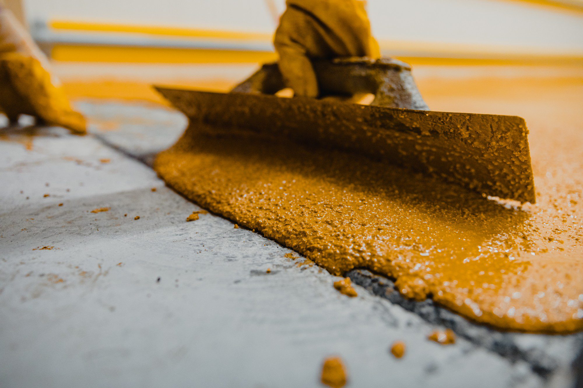 The image shows a close-up view of what appears to be a construction or industrial process. In the foreground, there is a large flat metal tool, potentially a floor scraper or squeegee, that is being used to spread or smooth a thick yellow substance on a concrete floor. The material being spread has a granular texture and golden colour, suggesting it might be some type of resin, epoxy, or other industrial coating.The person operating the tool is wearing protective gear, including gloves, and seems to be kneeling on the floor as they work. This type of activity is common in flooring applications where a uniform layer of a substance needs to be applied to a floor for sealing, leveling, or coating purposes.The background is out of focus, but we can see yellow lines, which might indicate caution areas or sections where work is being done or is yet to be done. The lighting is artificial, suggesting an indoor setting, possibly a factory, warehouse, or construction site.