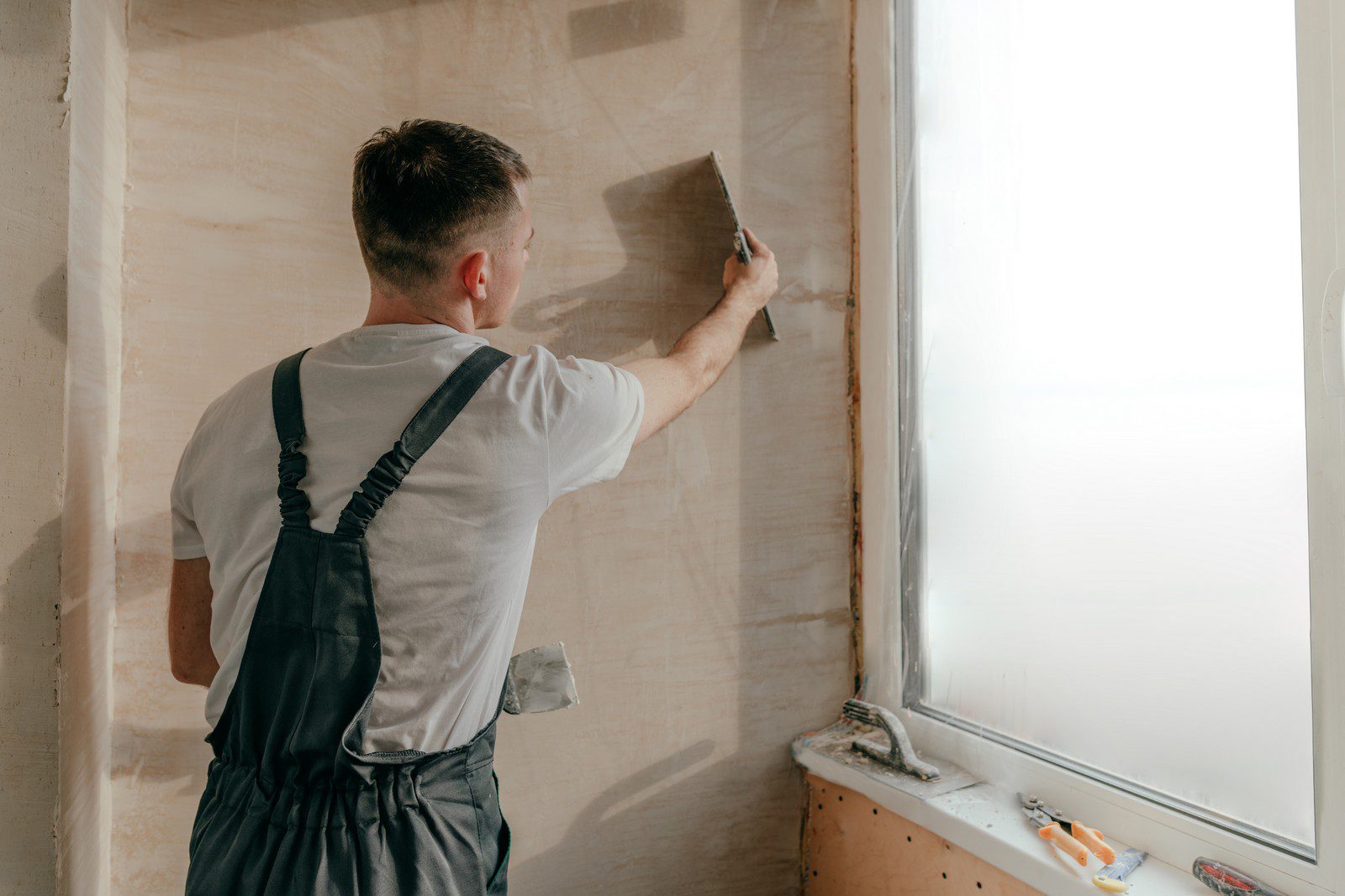 The image shows a person from behind engaged in a construction or home improvement task. They appear to be applying a compound to a wall using a taping knife, which suggests that they might be in the process of plastering or drywall finishing. The person is wearing work attire that includes a white T-shirt and dark-coloured overalls or braces with gray pants, typical of manual labour or a tradesman's uniform.The wall has a brown covering, which might be a layer of drywall compound that is being smoothed out. On the windowsill, there are additional tools consistent with drywall work, such as another taping knife and what looks like a drywall or putty knife, indicating that the person is in the midst of working.The room looks like it's under renovation with protective plastic covering part of the window to prevent splatters, and there's bright natural light coming in. The overall scene captures a moment of craftwork, focusing on the effort and skill involved in home renovation or construction.