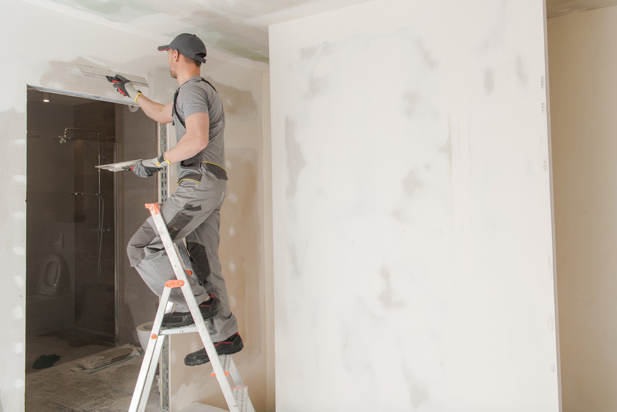 Certainly! Here is a detailed alt text description for the image at the provided URL:"A construction worker clad in a safety vest and a white hard hat is diligently patching a section of drywall. The worker uses a trowel to apply joint compound to a seam between two drywall panels. He is focused on smoothing the surface to create an even finish. The background reveals an unfinished interior space with exposed wooden framing and insulation visible. Various tools and materials are scattered on the floor nearby, suggesting an active construction site. The scene highlights the meticulous skill required in drywall finishing, with the worker carefully addressing imperfections to prepare the wall for the next stages of construction."