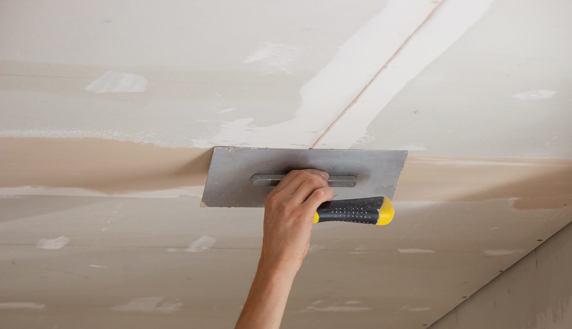 This image shows a person applying joint compound (also known as drywall mud) to a seam between sheets of drywall on a ceiling. The individual is using a wide drywall taping knife to smooth the compound over the taped seam, which is a step in the process of finishing drywall to create a smooth, paintable surface. The drywall has screw heads and tape already in place, and some areas have been previously mudded as part of the process.