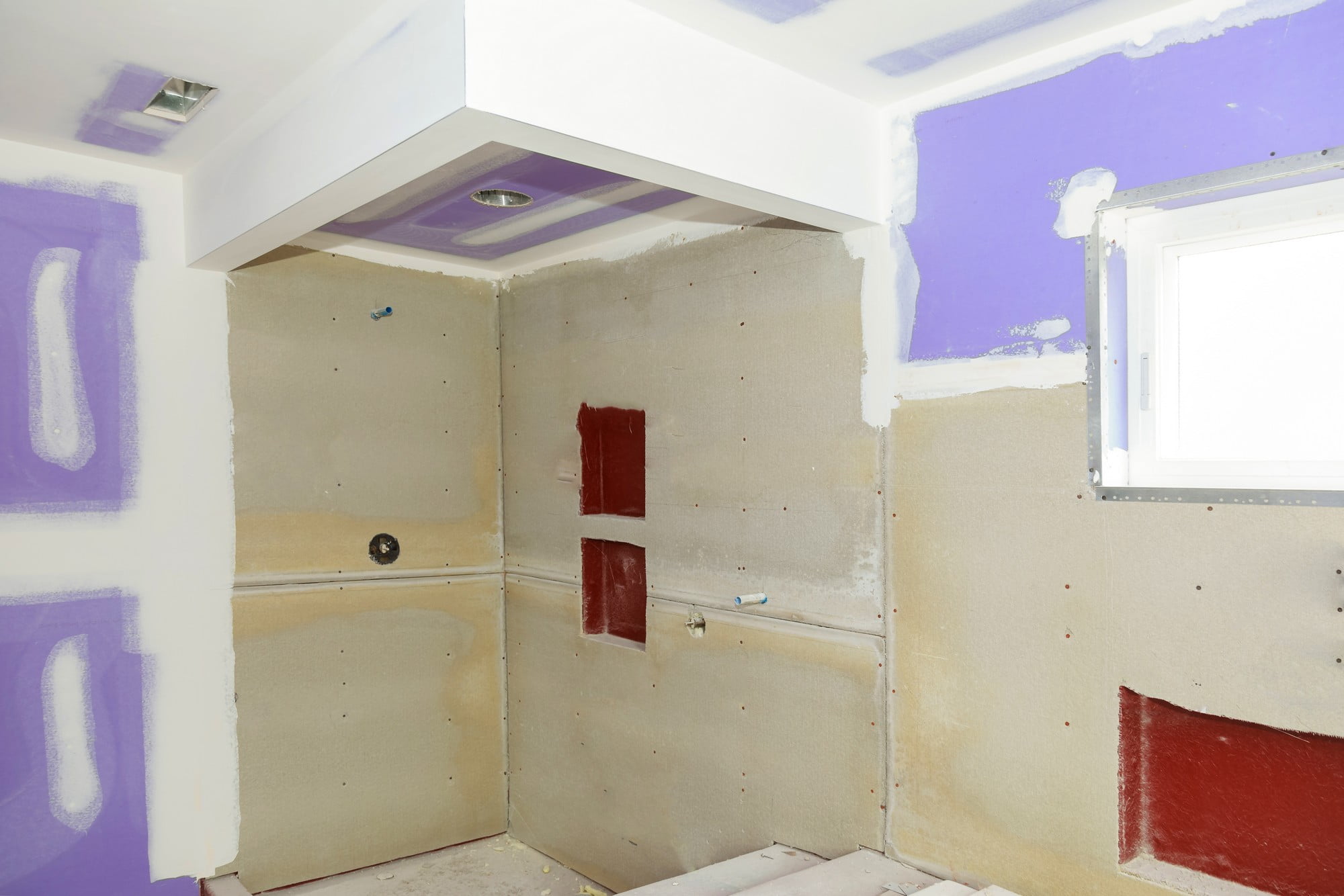 This image shows a bathroom undergoing renovation. You can see the walls are partially prepared with what appears to be cement board or drywall installed, some of which has been sealed with waterproofing membrane (the red areas), which is commonly used in wet areas to prevent moisture damage. There's visible purple drywall primer or compound on some sections of the wall, likely meant to indicate areas that need to be sanded or further prepared for tiling or finishing. The ceiling has recessed lighting installed, and a window is present to provide natural light. The floor is also not finished, suggesting that it might be prepared for tiling or another type of flooring soon. There are plumbing stub outs for a sink, and rough-ins for a shower or tub, indicating where fixtures will be installed. The unfinished state and visible construction materials indicate that the renovation is in progress.