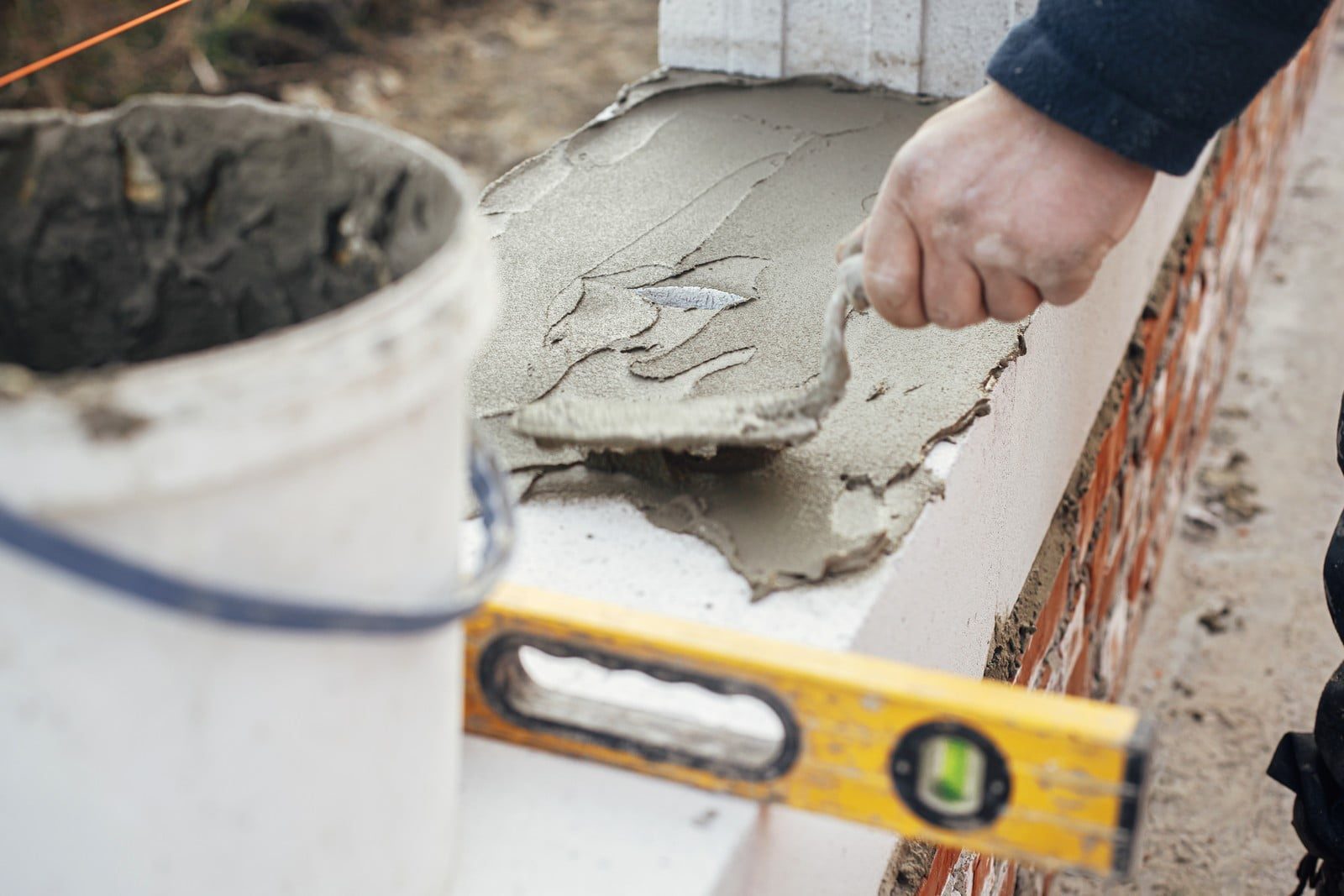 This image captures a construction or masonry process. A person's hand is shown applying wet cement or mortar on top of a concrete block using a trowel. In the background, there's a partially constructed brick wall. To the right, a spirit level is resting on the concrete block, likely for ensuring the structure is level. On the left side, you can see a bucket that appears to contain more of the cement or mortar mix. The overall scene suggests masonry work being done, possibly as part of building a new structure or repairing an existing one.