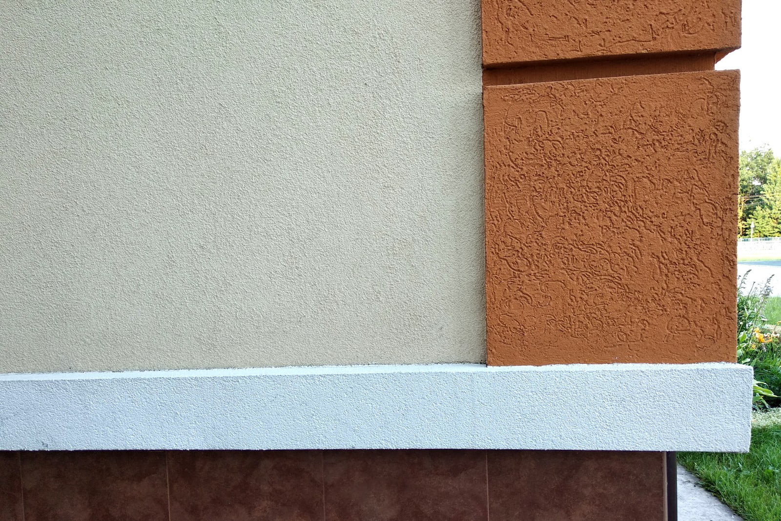 This image shows a portion of a building's exterior wall. On the bottom, there is a section of brown tiles that form a baseboard-like feature along the ground. The main part of the wall is painted in a light beige colour with a textured finish. On the right side, there is an architectural feature that protrudes from the wall; it's painted in a darker, rust-like colour, also with a textured finish. This could be a decorative column or pilaster. The corner where these two wall surfaces meet has a sharp, 90-degree angle delineating the change in colour and texture. On top of the brown tiles, there is a white styrofoam insulating material which provides a contrast and possibly serves as insulation or a decorative mould. There is a grassy area visible in the background reflecting that the building is located in a green outdoor area.