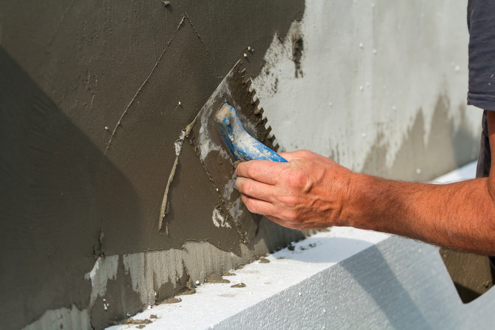 This image shows a close-up of a person's hands applying wet cement or plaster to a vertical surface using a notched trowel. The hands are partially covered in the material, and the individual seems to be in the process of smoothing it out or spreading it on the wall, which is a common step in construction or masonry work when creating a smooth, flat surface for tiling, rendering, or plastering.