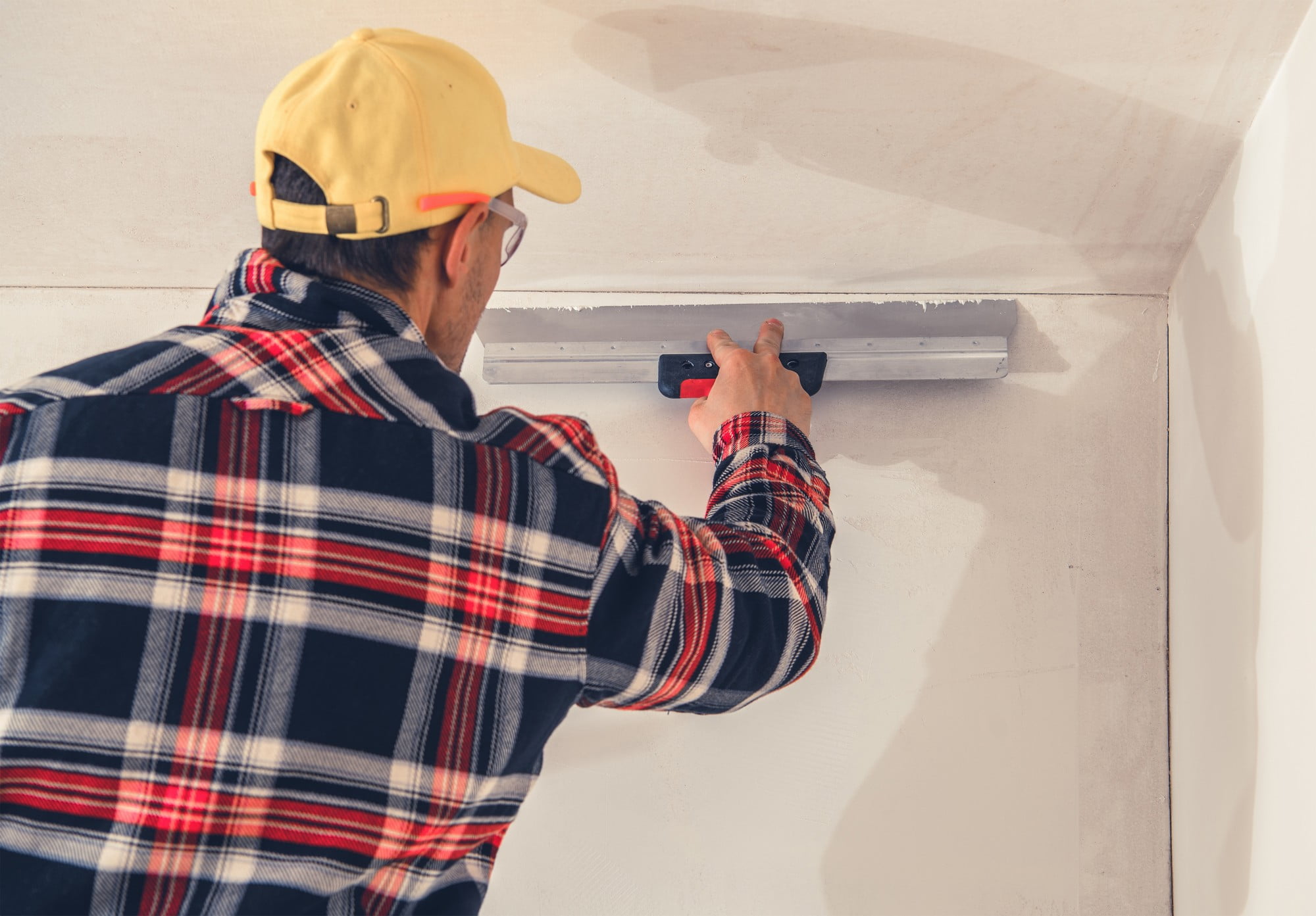 The image shows a person applying a finishing touch to a wall. They are using a plastering trowel to smooth out the surface of what appears to be a freshly applied layer of plaster or joint compound. The individual is wearing glasses, a yellow cap, and a plaid shirt. The environment suggests construction or renovation work being done indoors.