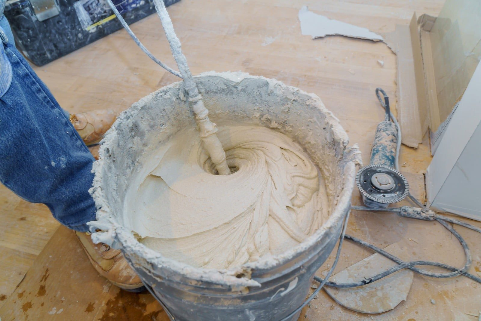 The image shows a close-up view of a construction or renovation scene. There is a plastic bucket filled with mixed joint compound or plaster, looking creamy and thick with swirling patterns, which indicates that it has recently been stirred or mixed. Partially visible is the lower part of a person wearing blue denim and work boots, standing next to the bucket, suggesting they are the person doing the work.There's a mixing tool or paddle attached to a drill protruding from the joint compound, covered in the mixture, which has been used to mix the compound. We can also see a construction tool with a circular blade, possibly used for cutting drywall or similar materials, lying on the floor. There are bits of debris and some work equipment scattered around the scene. The floor is covered in protective material, likely to prevent damage or mess from the construction work.