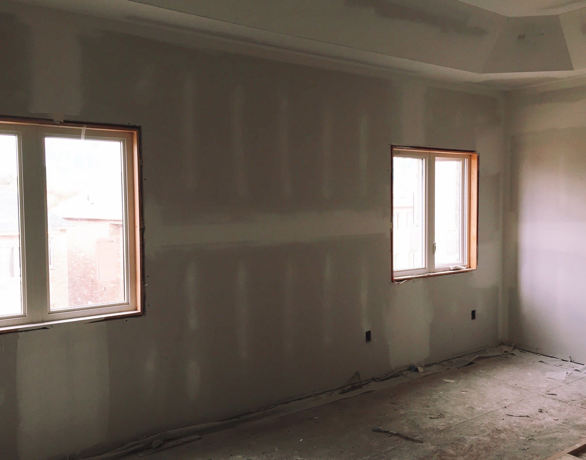 The image shows an interior room under renovation or construction. The walls are unfinished with visible drywall and joint compound. There are two windows that provide natural light, and the ceiling has a recessed, tray-like feature often referred to as a tray ceiling. There is construction debris on the floor, and electrical outlets are visible without cover plates, indicating ongoing work. The space is empty, suggesting that the room is in a transitional phase, possibly being prepared for painting, flooring, or other finishing work.