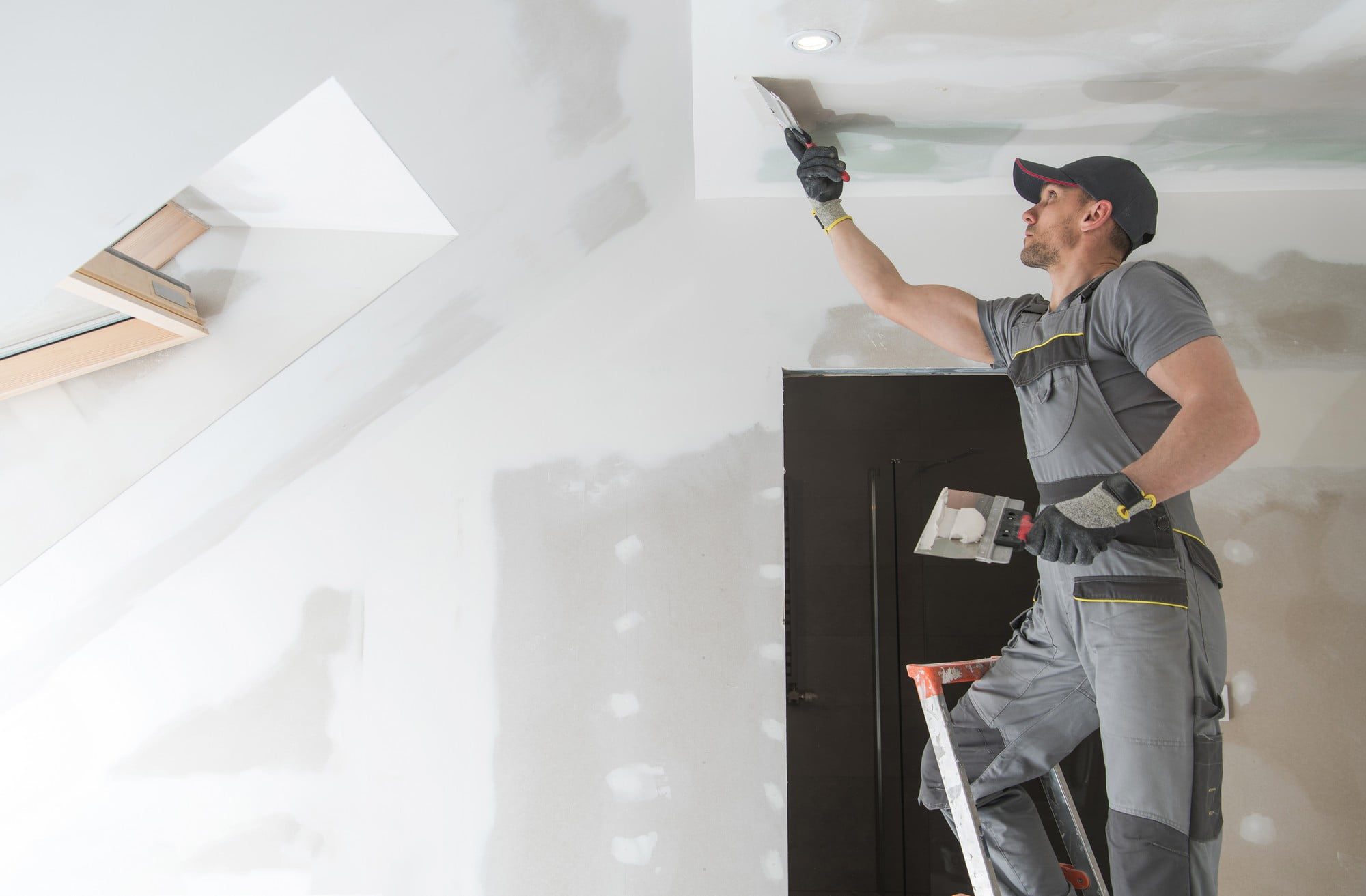 The image shows a person working on the interior finishing of a room. The worker is standing on a stepladder and appears to be applying joint compound, also known as drywall mud, with a taping knife to the seams of drywall panels on the ceiling. The work is part of the drywall installation process, which involves smoothing over the joints and screws to prepare the walls for painting or wallpaper. The room is unfurnished, and several patches of drywall mud can be seen applied on the walls and ceiling, indicating that the finishing work is in progress. Additionally, there's a skylight installed in the ceiling, providing natural light to the space. The worker is wearing a gray work outfit with reflective safety detailing, a cap, and gloves, indicating adherence to safety and workwear standards. It looks like a professional construction or renovation scene.